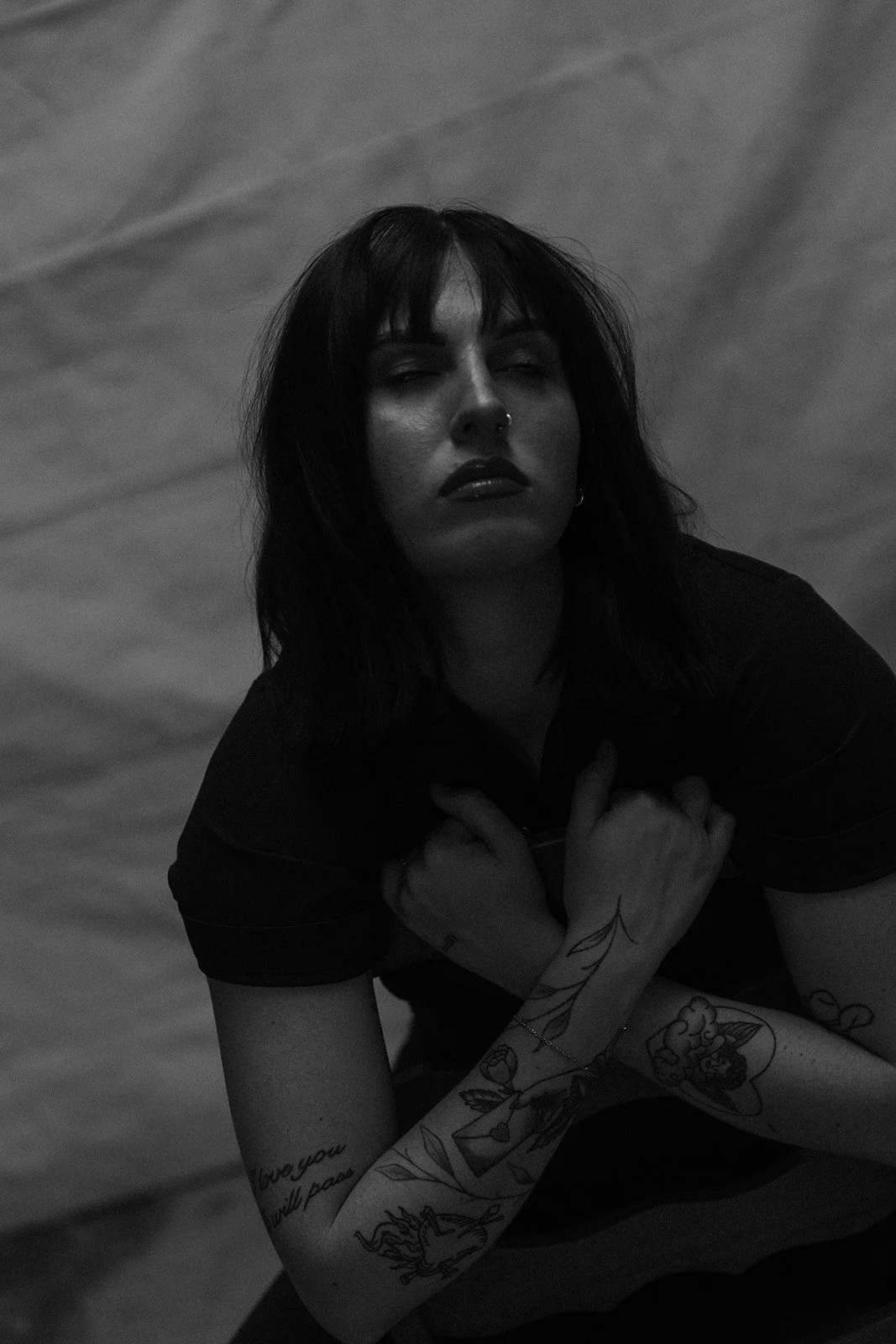 A woman with dark hair and tattoos on her arms, wearing dark clothing, poses with her hands crossed over her chest, eyes partially closed, against a slightly blurred background.