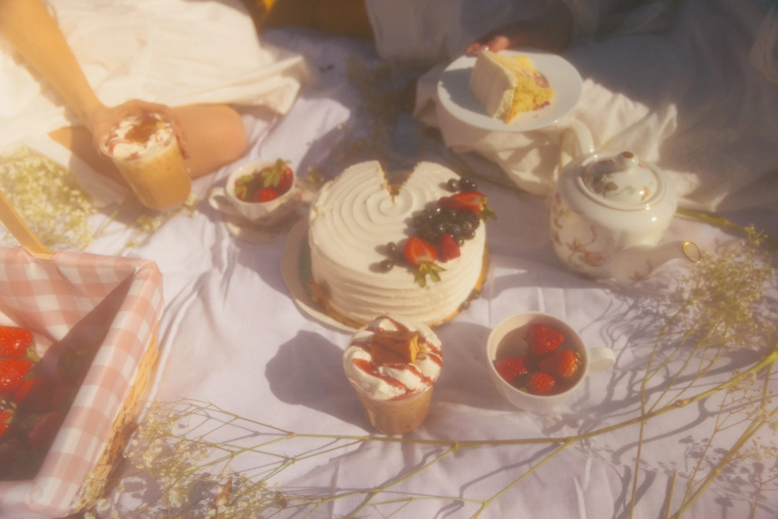A variety of cakes and desserts on a table, including a layered cake with strawberries and blueberries, small cups of strawberries, a cake roll, and a teapot, with a basket and other tableware nearby.
