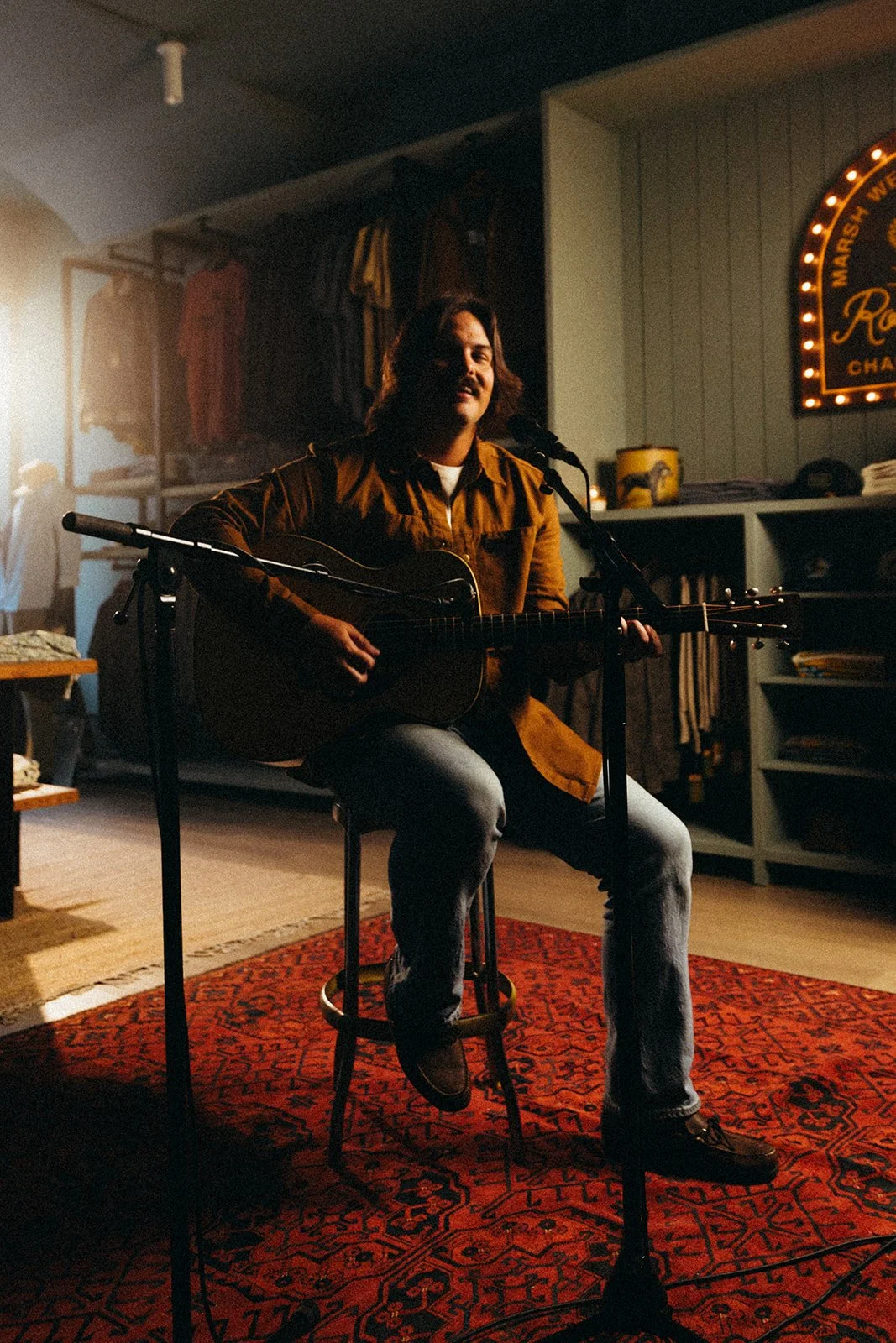 A man with long hair and a beard sitting on a stool, playing an acoustic guitar and singing into a microphone in a cozy, dimly lit room, possibly a music store or studio, with colorful clothing hanging on the wall and a neon sign in the background.