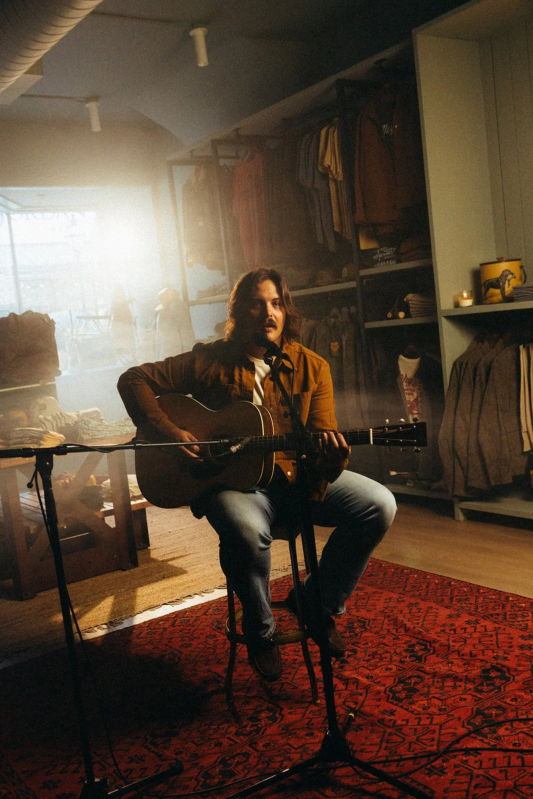 A man with shoulder-length hair sitting on a chair playing an acoustic guitar, inside a store with shelves of clothing and items, with sunlight streaming in from a window behind him.
