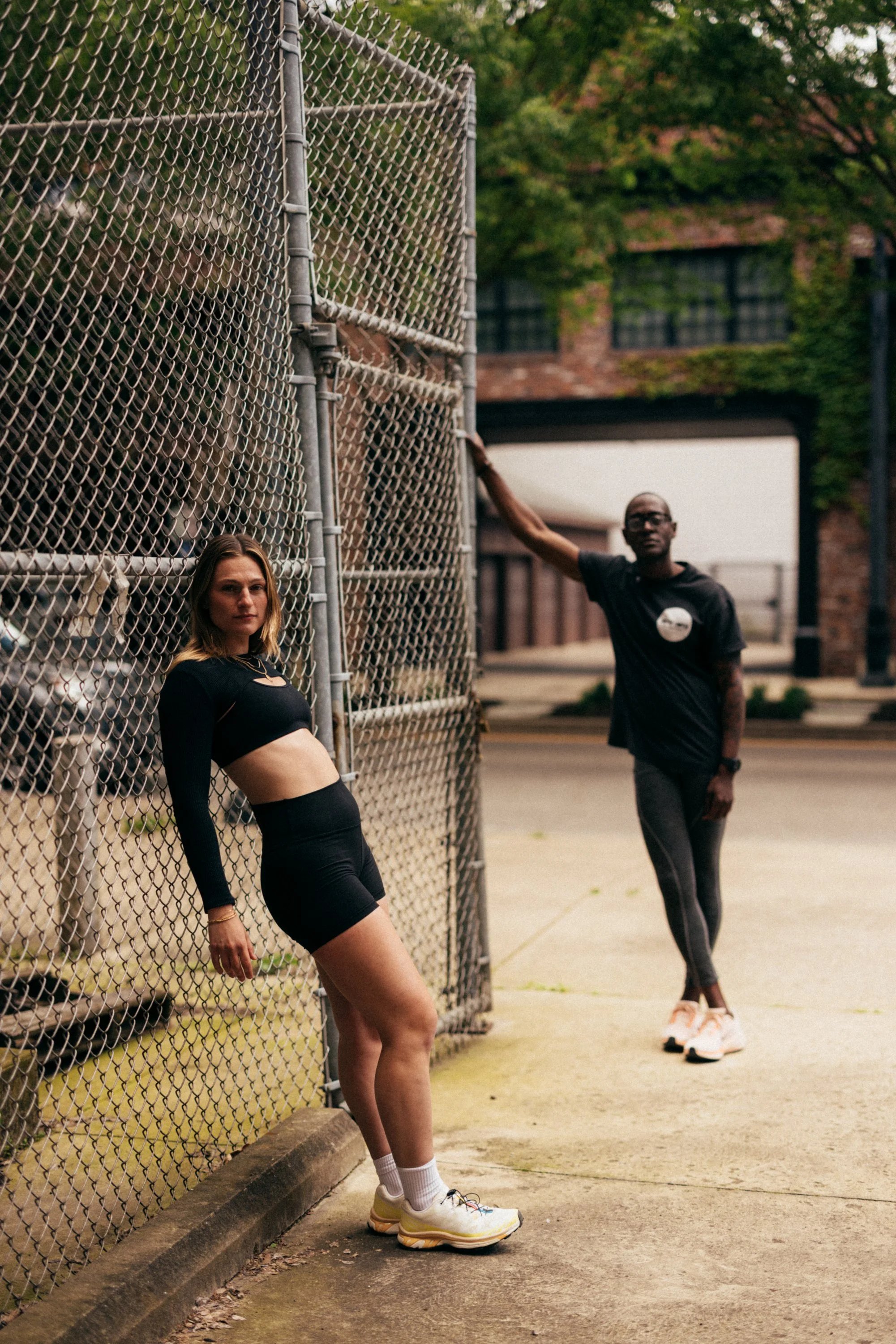 A young woman leaning against a chain-link fence on a city sidewalk, with a man standing in the background near a brick building with green foliage.
