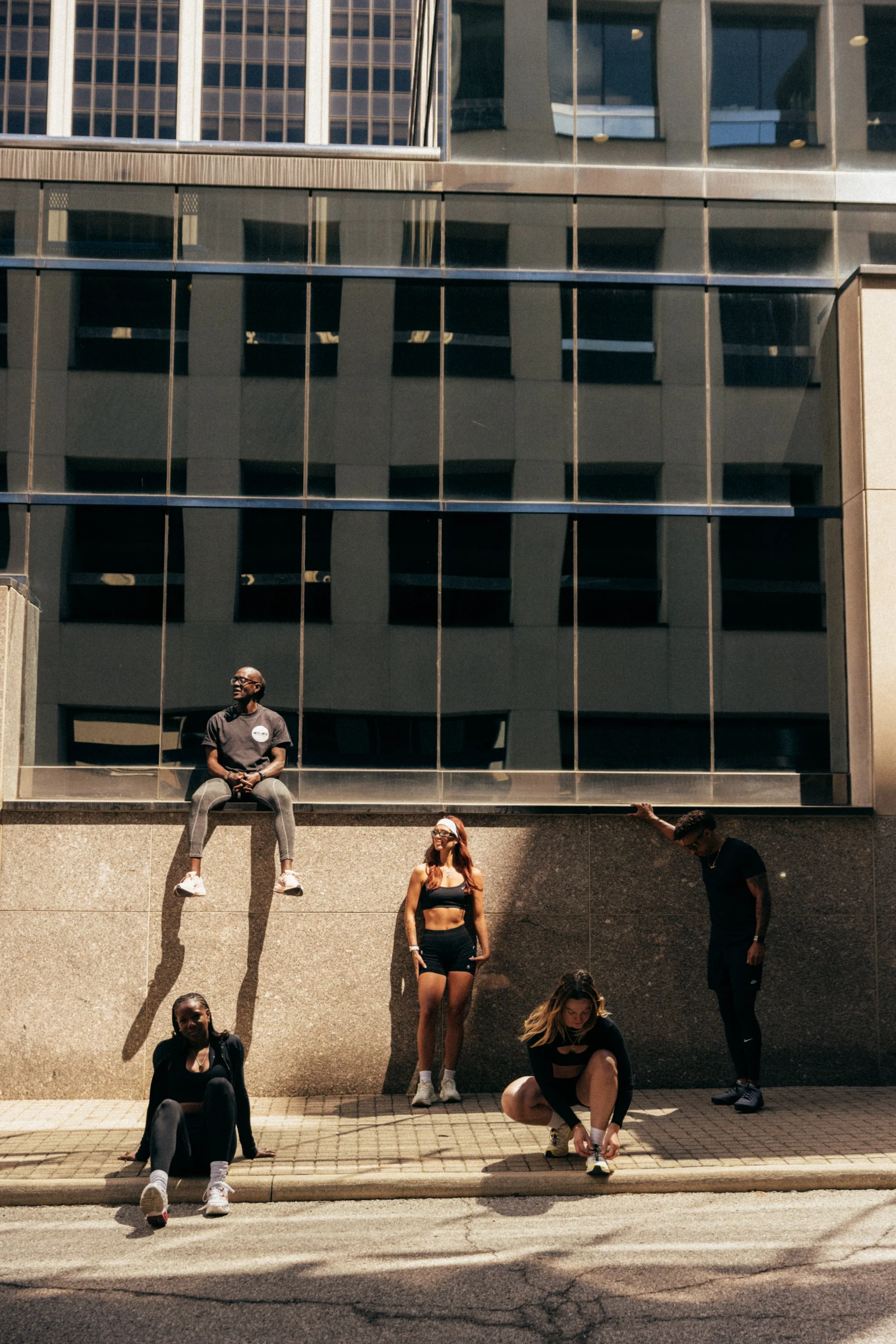 People sitting and standing in front of a glass building, with some individuals tying shoes and others posing or resting.