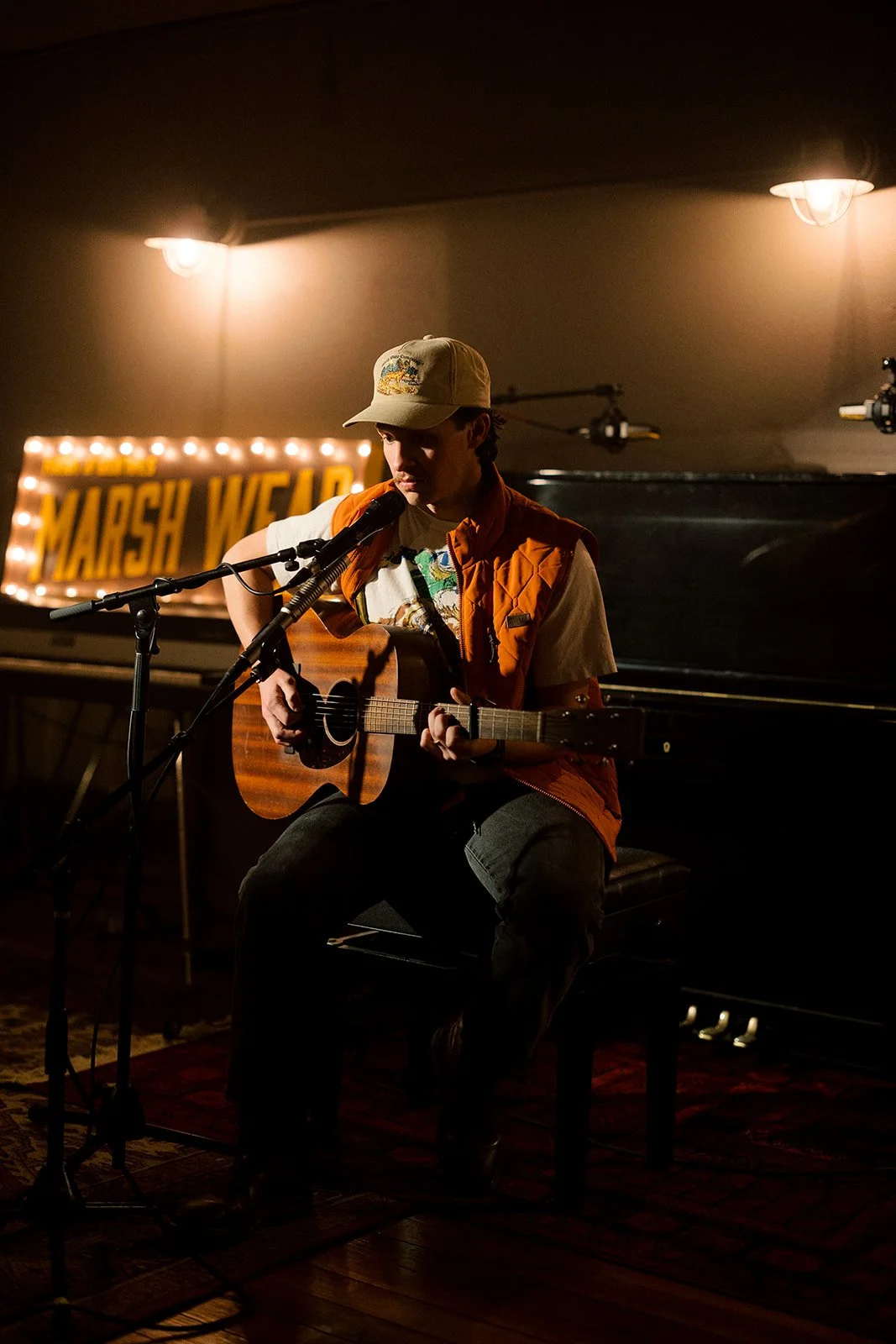A person wearing a beige cap and orange vest plays an acoustic guitar on stage, sitting on a black bench, in front of a microphone, with a illuminated sign reading 'Marsh W' in the background.