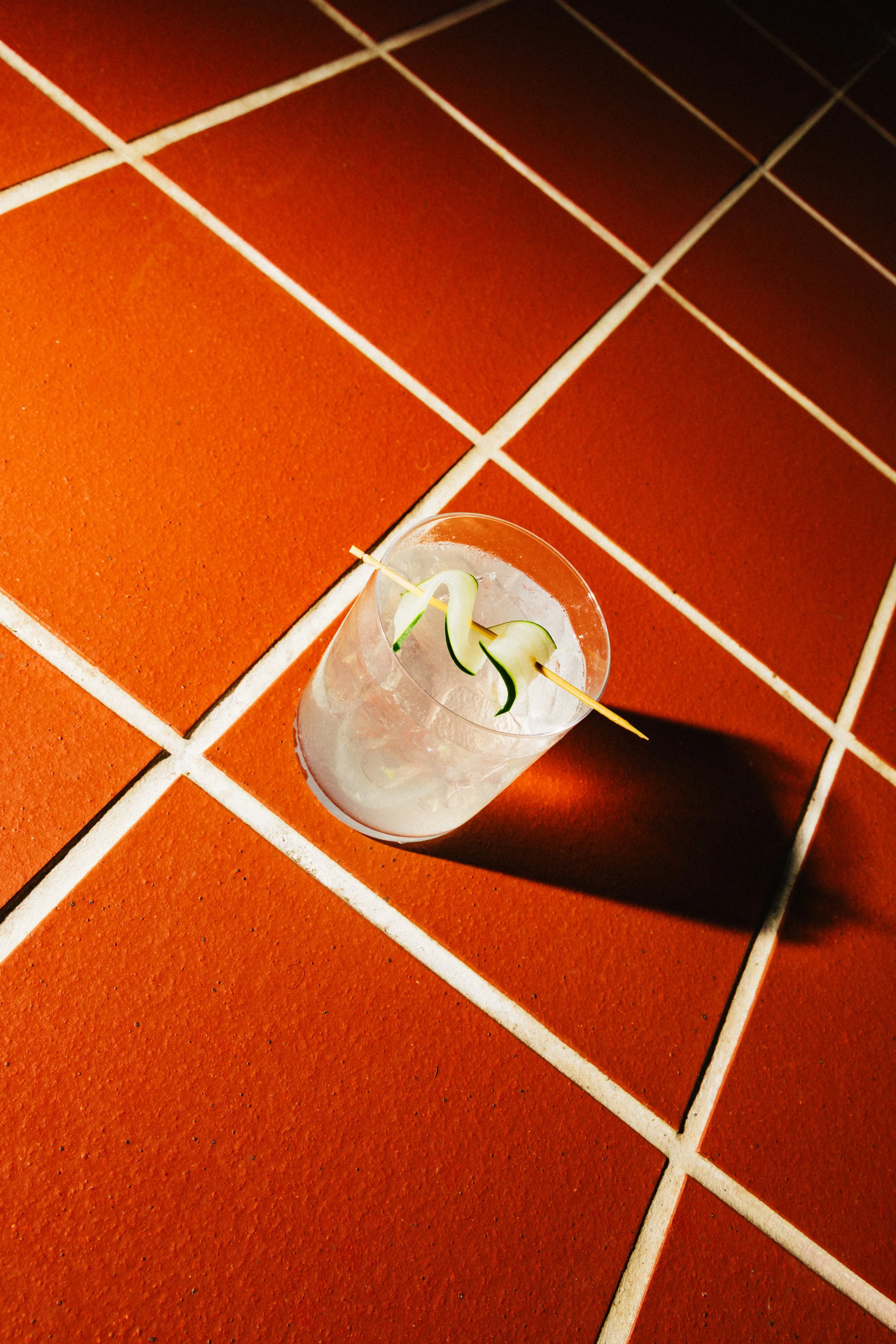 A clear glass with a cocktail garnished with a thin slice of cucumber, placed on a terracotta tiled floor.