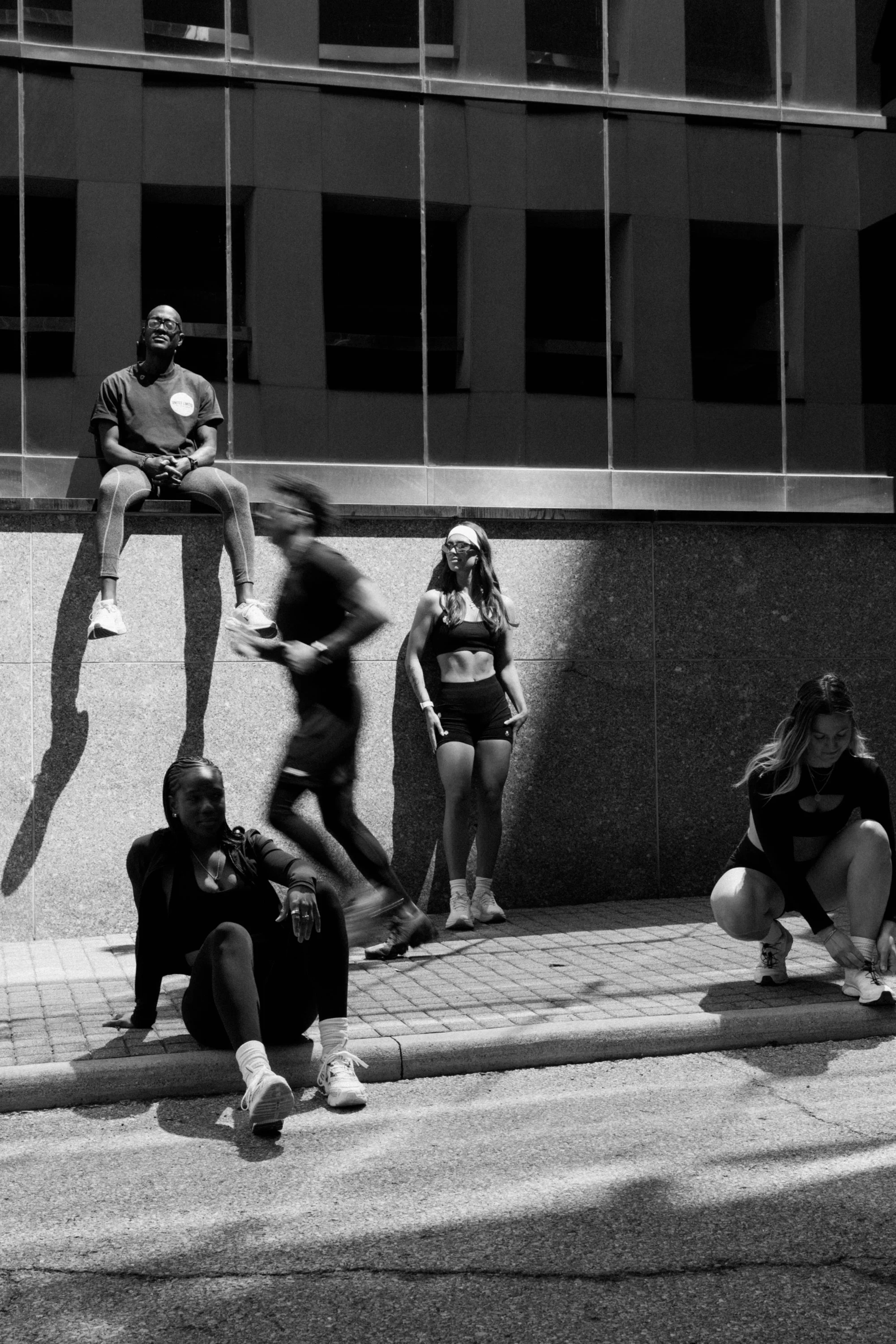 Black and white photo of six young people outdoors in front of a modern building. Two are sitting on the ground, one is standing, one is climbing, and two are sitting on a ledge. The people are casually dressed, and some are wearing glasses.