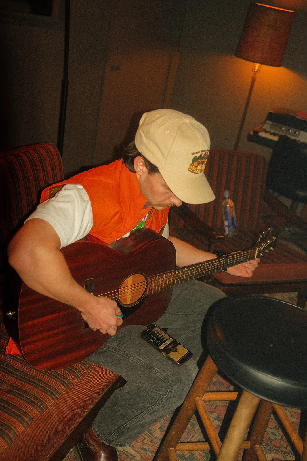 A man wearing a beige cap and orange vest playing a guitar in a dimly lit room.