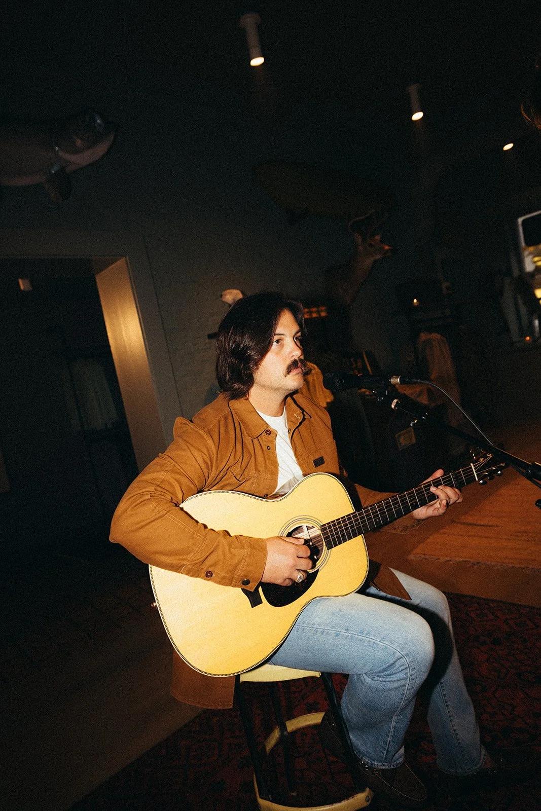 A man with dark hair and a mustache is sitting on a stool, playing an acoustic guitar and singing into a microphone in a dimly lit room.