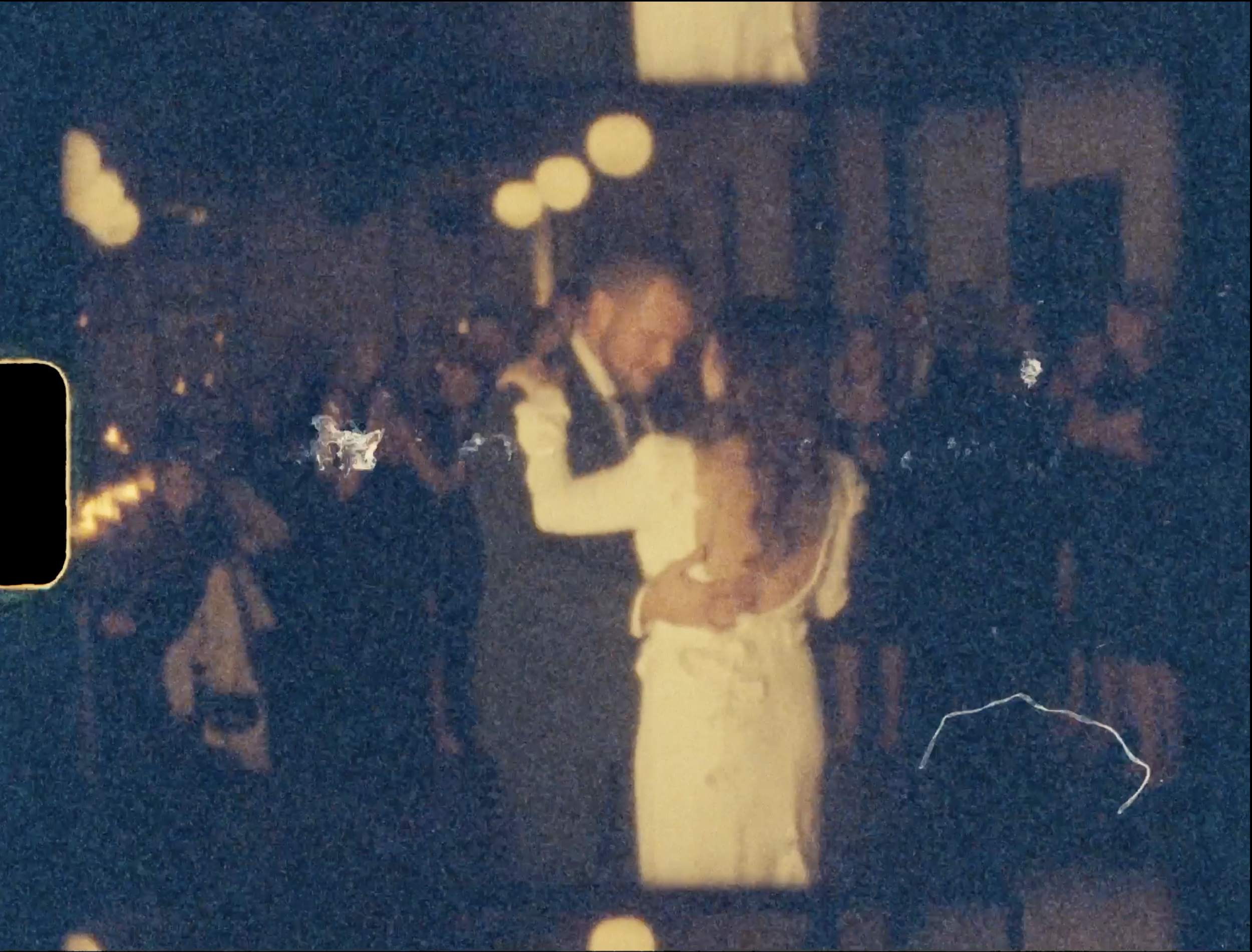 A man and a woman are dancing closely at a nighttime event. The man has short dark hair and is wearing a dark jacket, while the woman has long hair and is wearing a light-colored dress. The background shows other people sitting at tables in a dimly l