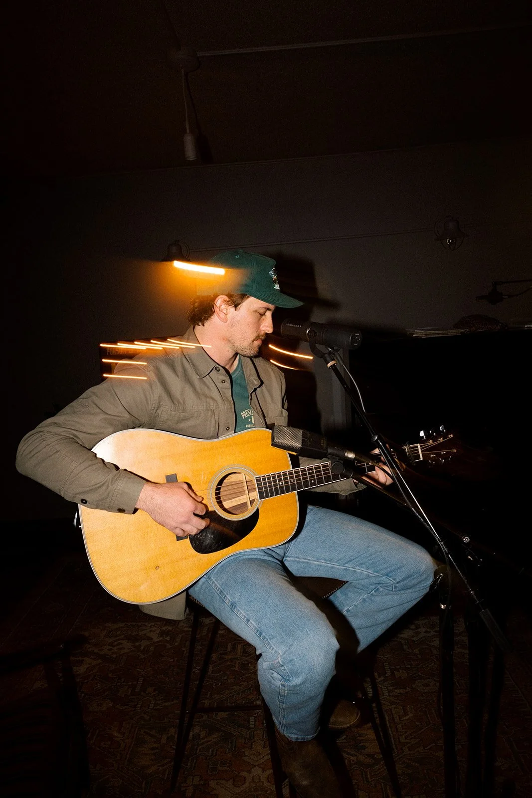 A man sitting on a chair, playing an acoustic guitar and singing into a microphone in a dimly lit room, with light streaks indicating movement or camera exposure effects.