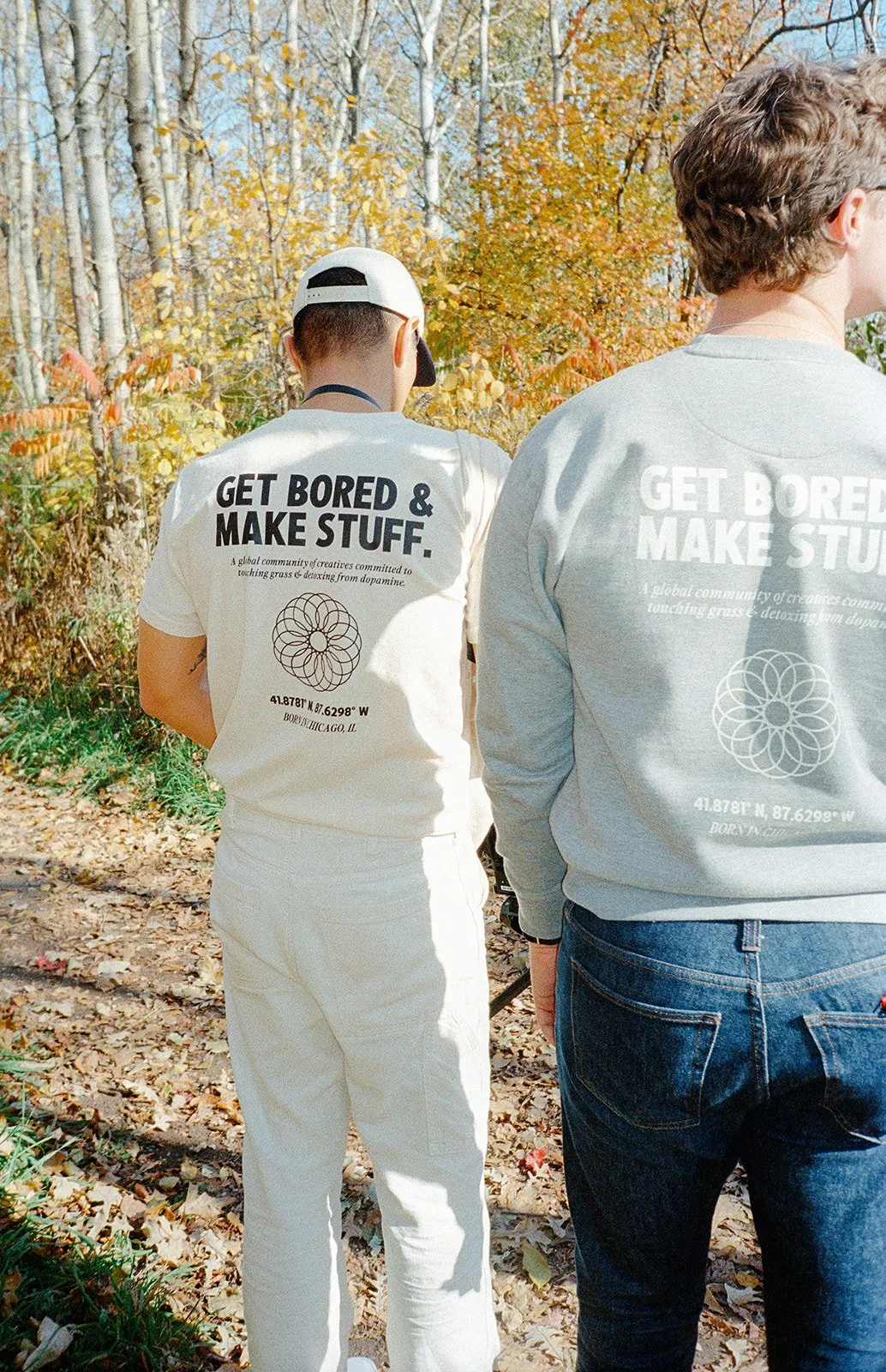 Two people standing outdoors in fall clothing, facing away, with trees and fallen leaves around them.