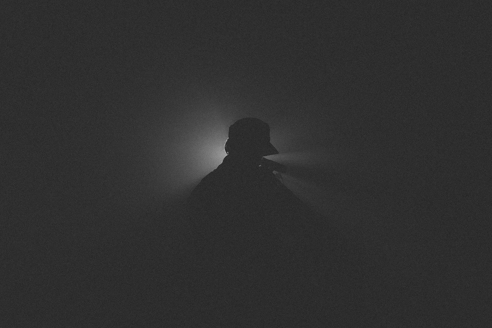 Person wearing a hat and jacket, standing in front of a light source behind them in a dark, foggy environment.