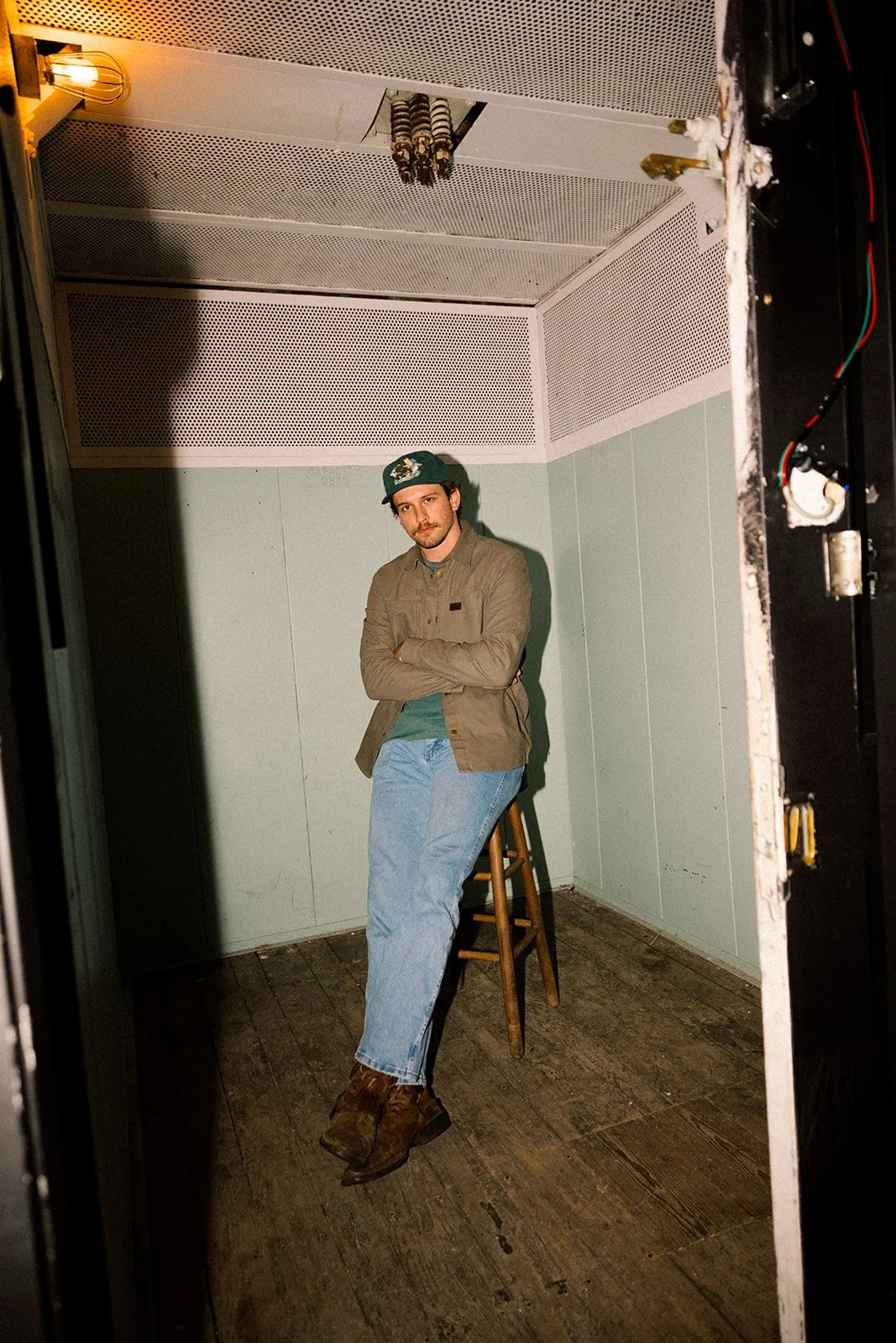 A man with dark hair and a mustache, wearing a green cap, tan jacket, blue jeans, and brown boots, is sitting on a stool inside a small room with plain light-colored walls and a wooden floor, looking at the camera with arms crossed.