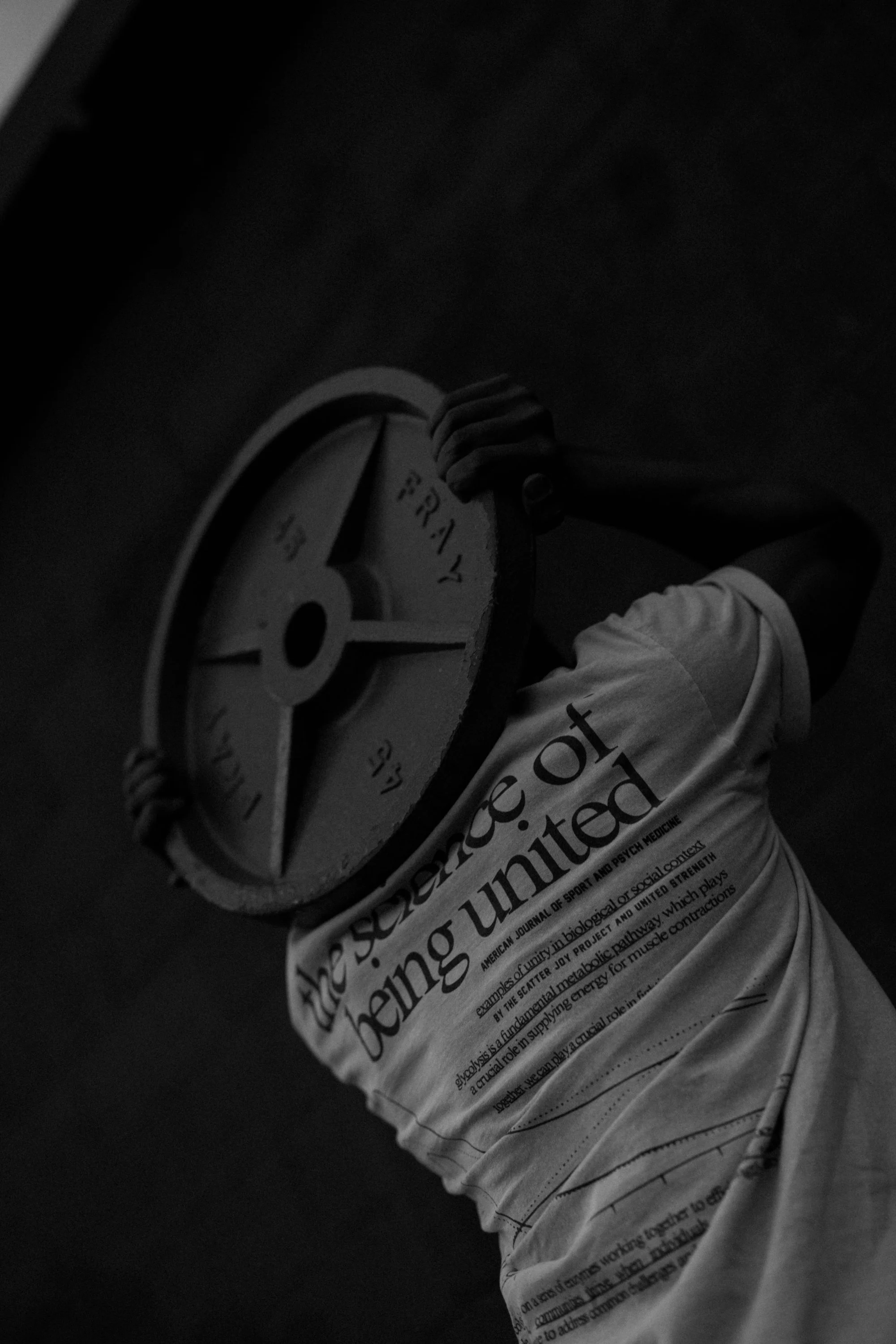 Person lifting a barbell in a gym, wearing a T-shirt with text, with a newspaper in the foreground.