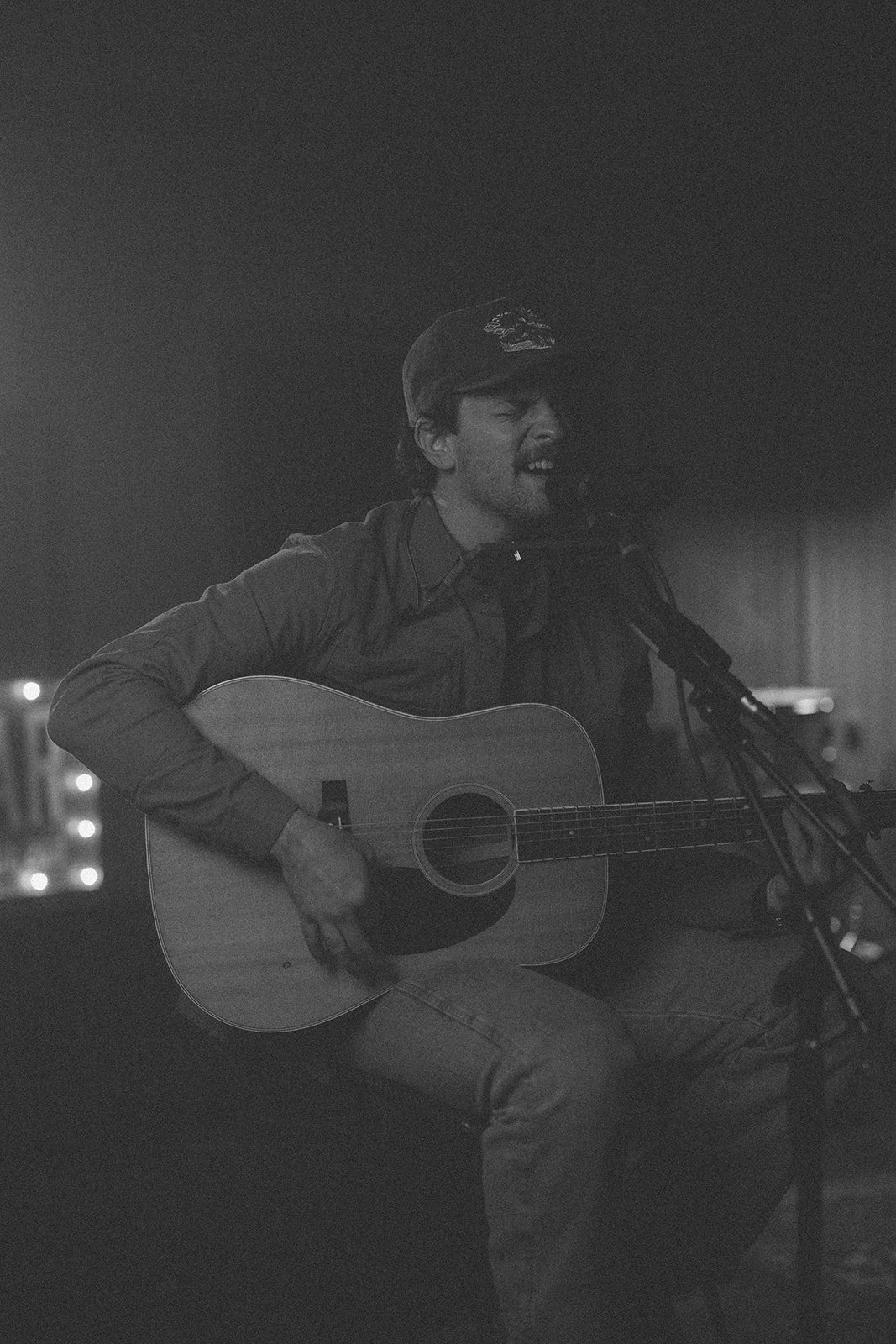 A man wearing a cap sings into a microphone while playing an acoustic guitar in a dimly lit setting, possibly a stage or recording studio, in black and white.