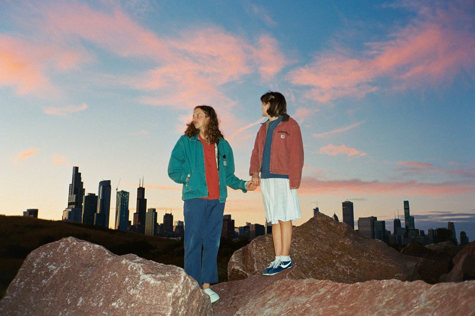 Two people holding hands on large rocks during sunset with the Chicago skyline in the background.
