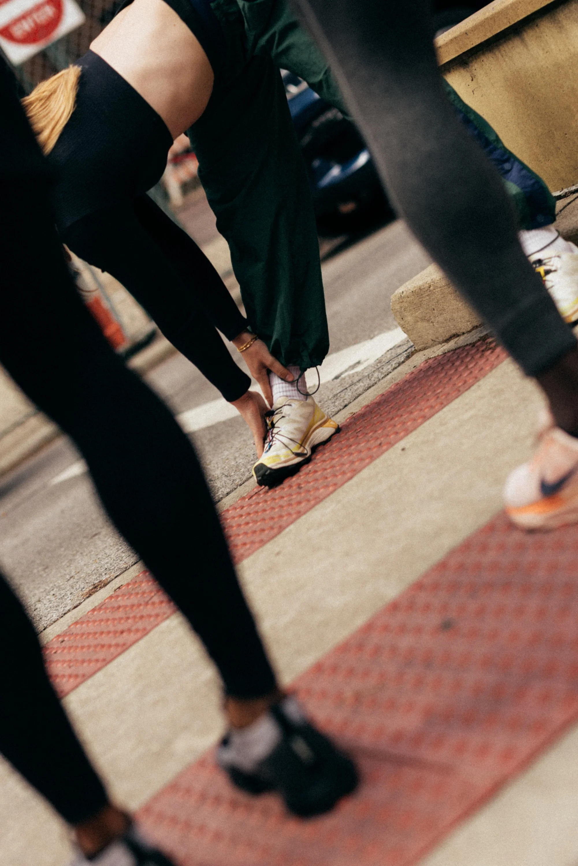 A person sitting on a yellow stool, tying the shoelaces of their white and yellow athletic shoes on a city street sidewalk, surrounded by a black railing and a red tactile paving strip.