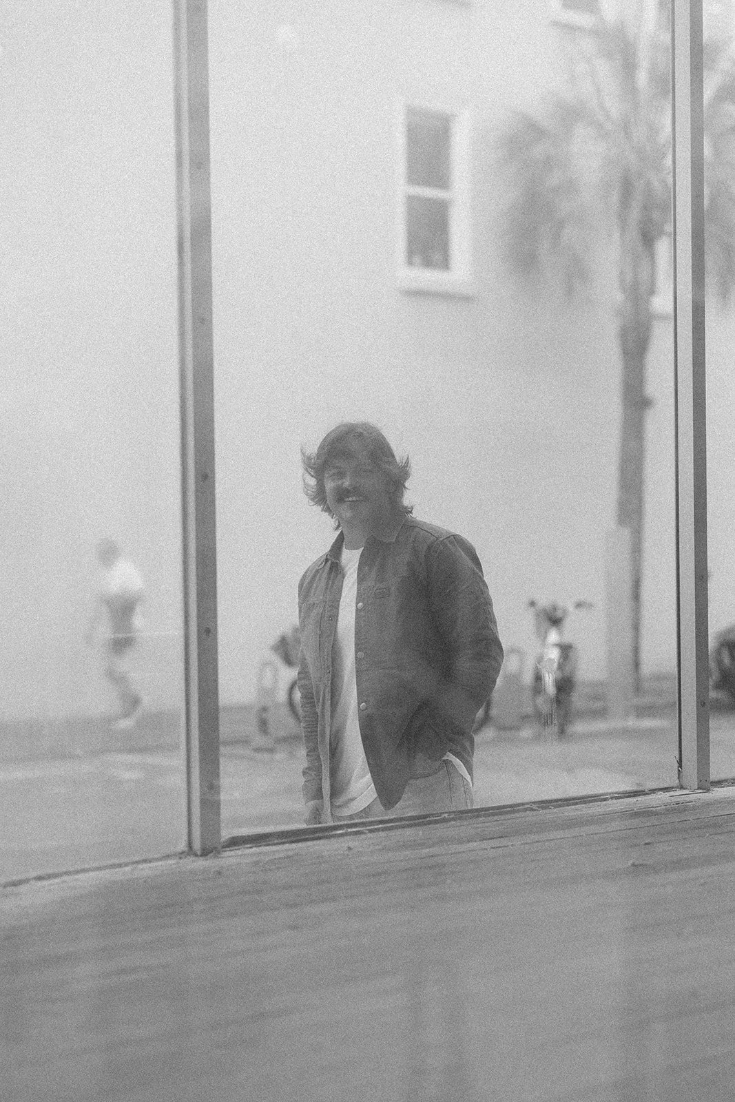 A young man with long hair and a beard, smiling and standing outside behind a window, wearing a jacket and a t-shirt, with blurred bicycles and trees in the background.