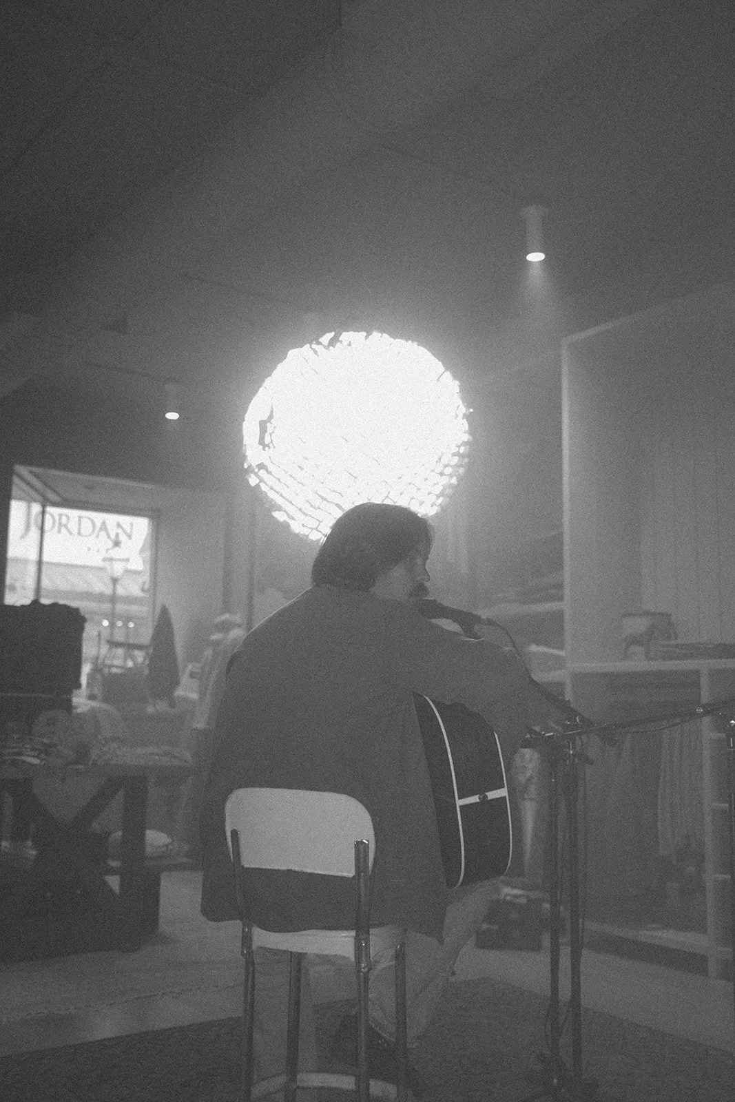 A person playing guitar and singing into a microphone, seated on a chair, in a dimly lit indoor setting with bright circular window or light source behind.
