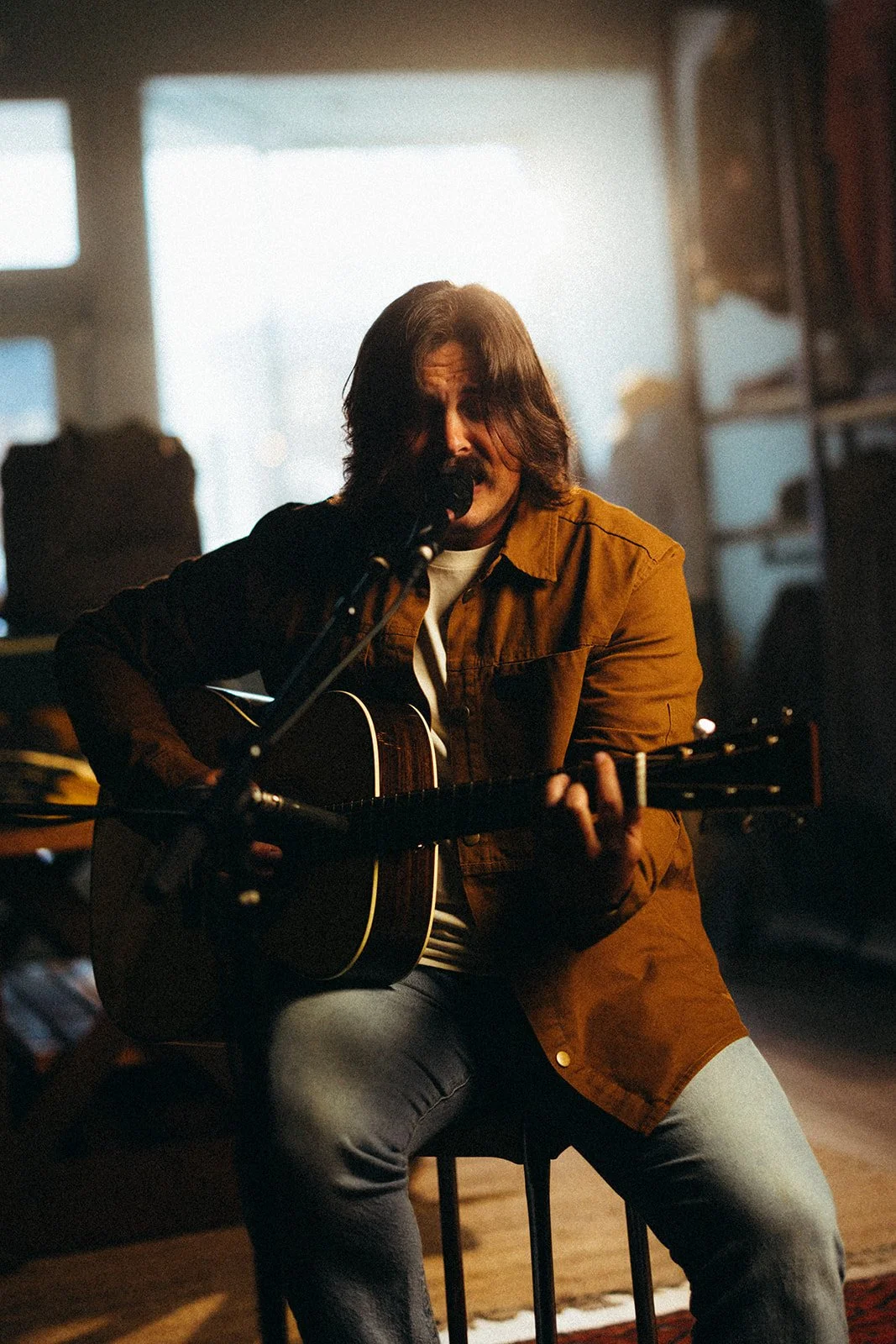 Man with long hair and mustache playing acoustic guitar and singing into a microphone in a cozy room with warm lighting.