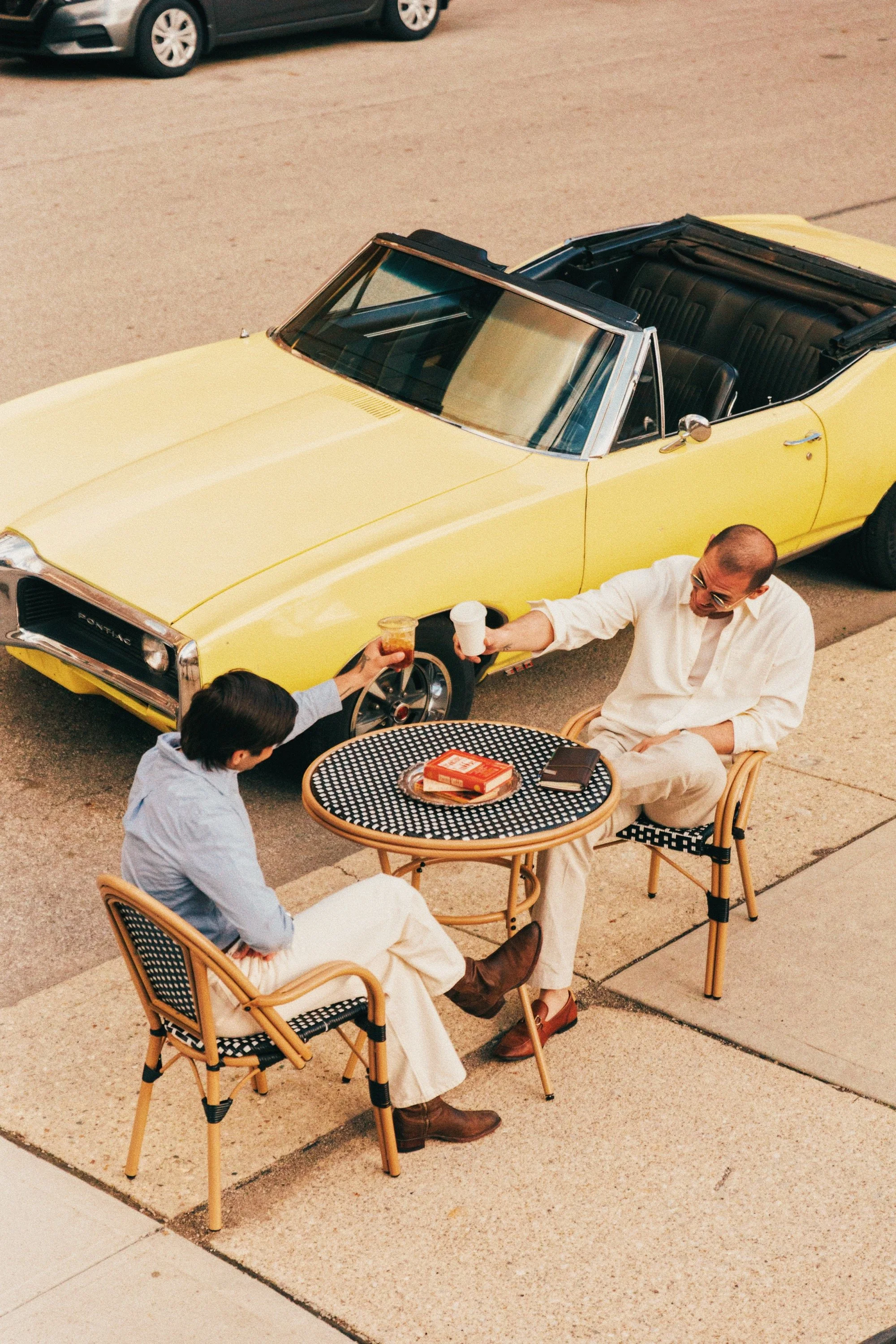 Two men sitting at outdoor tables exchanging drinks near a yellow convertible car parked on a paved surface.