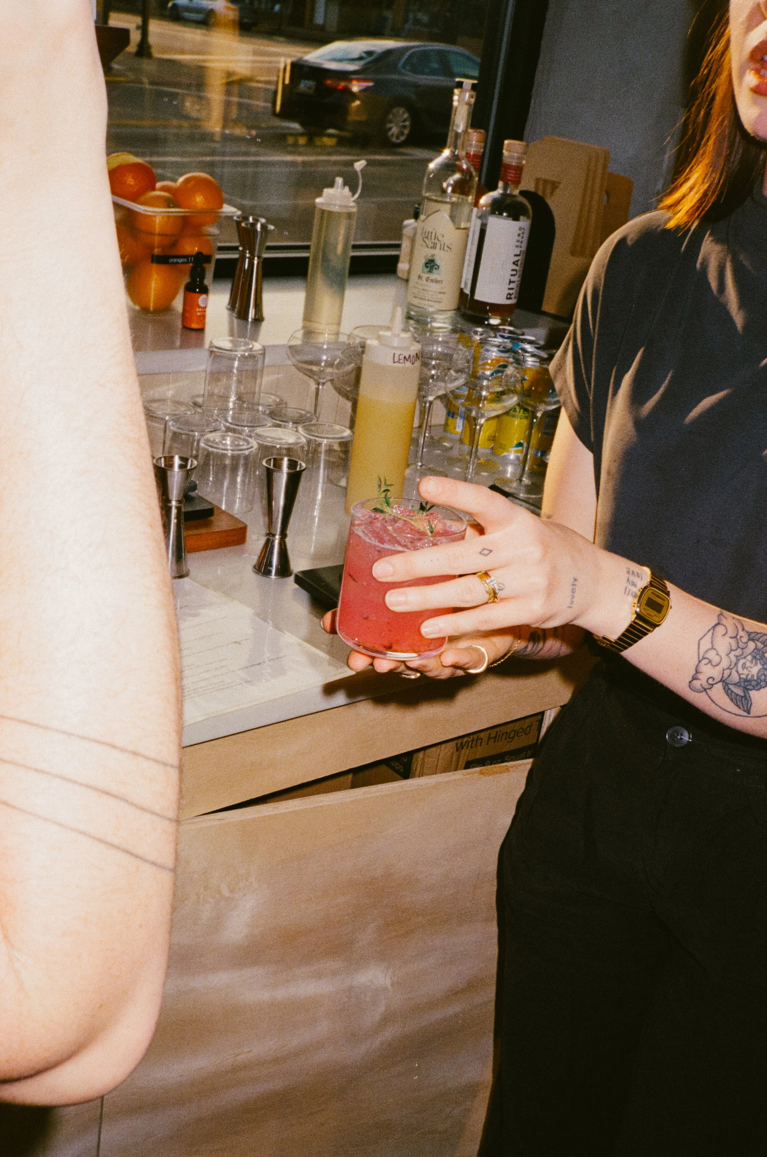A person holding a pink cocktail with a garnish, standing at a bar with various bottles and bar tools, and another person's arm visible in the foreground.