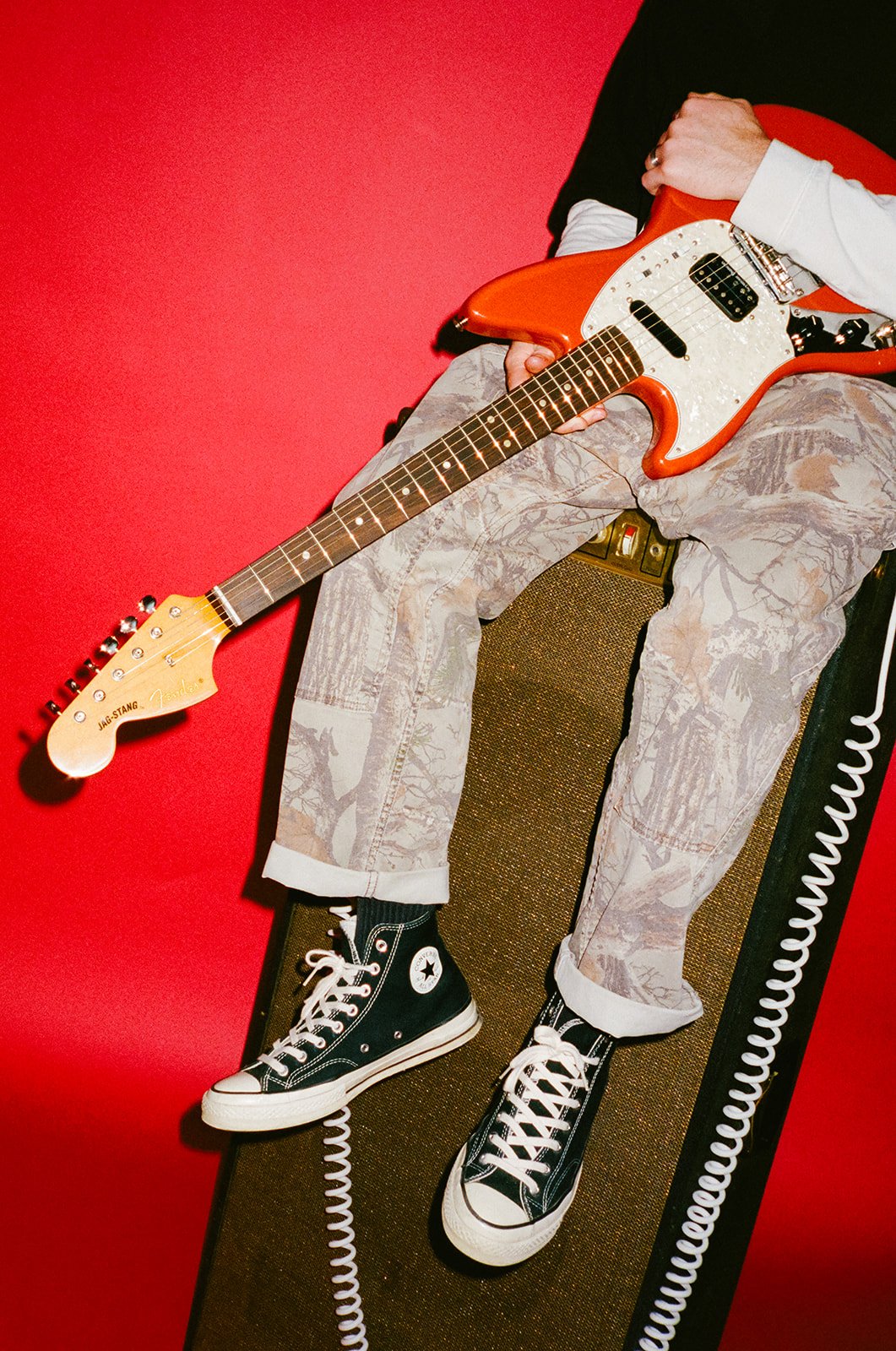 A person sitting on a vintage amplifier holding an orange electric guitar against a red background. The person is wearing camouflage pants, black high-top Converse sneakers, and a white long-sleeve shirt.