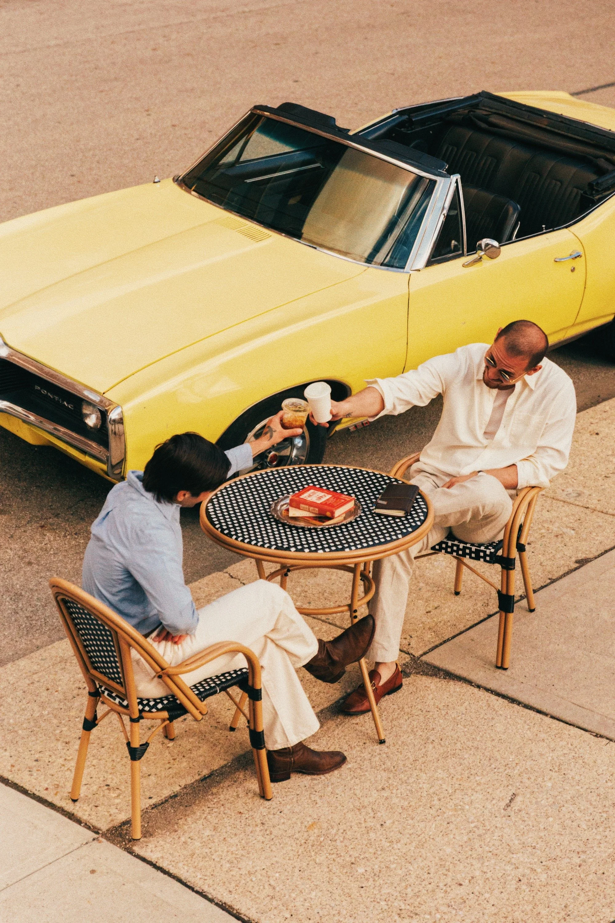 Two men are sitting at a black and white checkered outdoor table on a sidewalk, having drinks. One man in a beige outfit and sunglasses is smiling, while the other man, wearing a light blue shirt, is reaching for a drink. Cafe Stai Manso