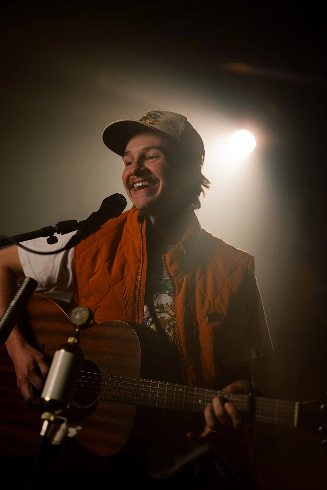 A young man performing with a guitar and microphone, smiling, wearing a cap and an orange vest, with a bright light behind him.