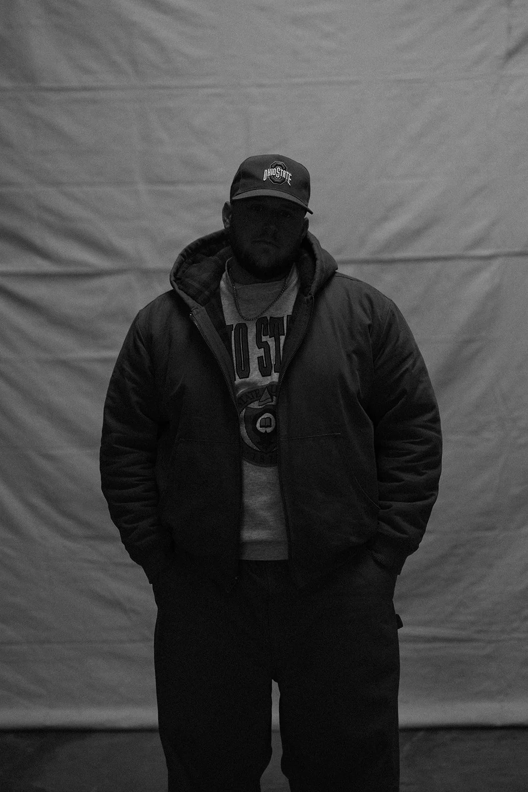 A man standing against a plain backdrop wearing a baseball cap with 'Ohio State' logo, a hoodie under a jacket, and dark pants, with one hand in his pocket.