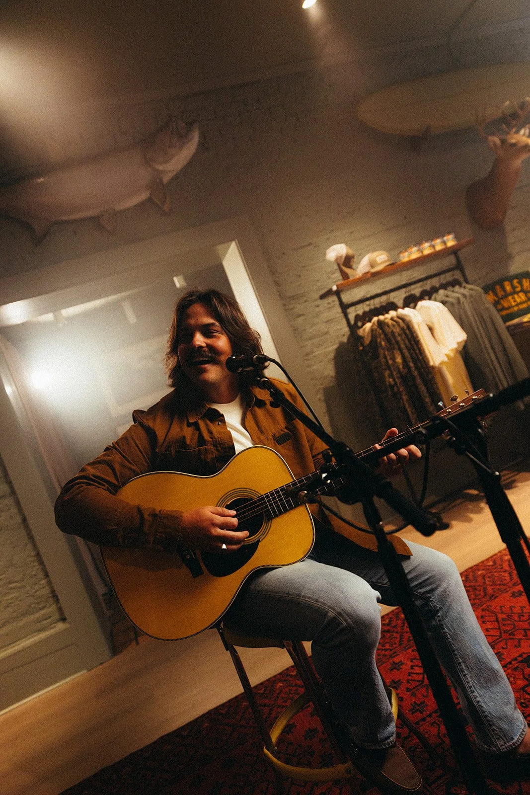 A man playing an acoustic guitar and singing into a microphone in a cozy indoor setting with wooden flooring, a red patterned rug, and shelves with clothing and decorative items in the background.