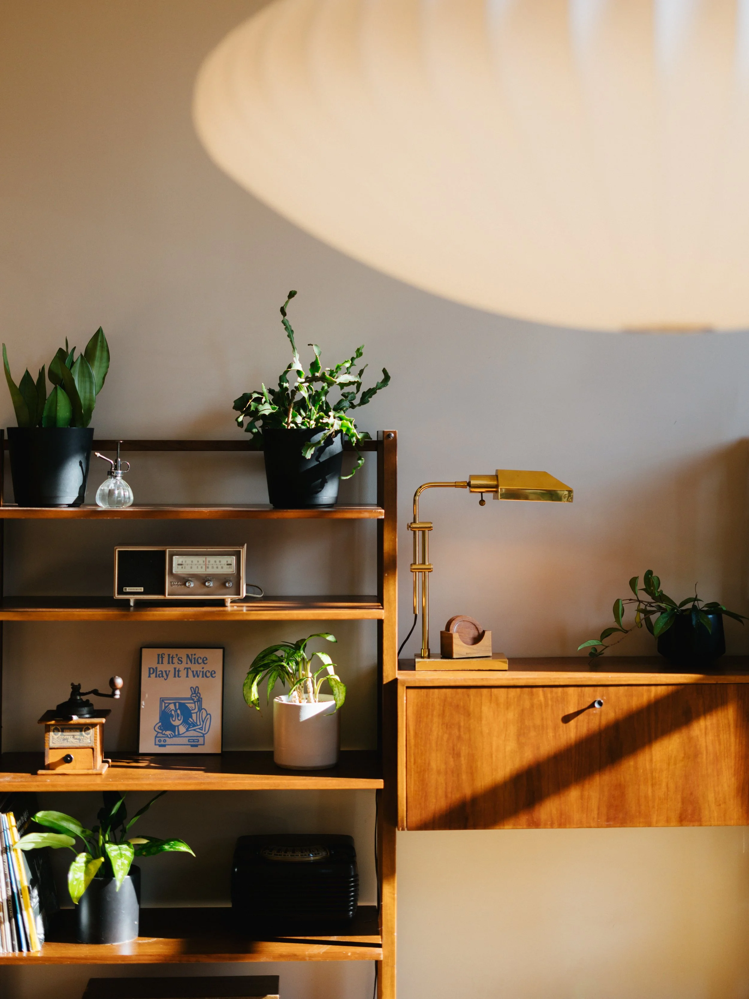 A wooden bookshelf with plants, a vintage stereo, a framed quote, and a brass desk lamp, illuminated by soft sunlight and a large white ceiling light.