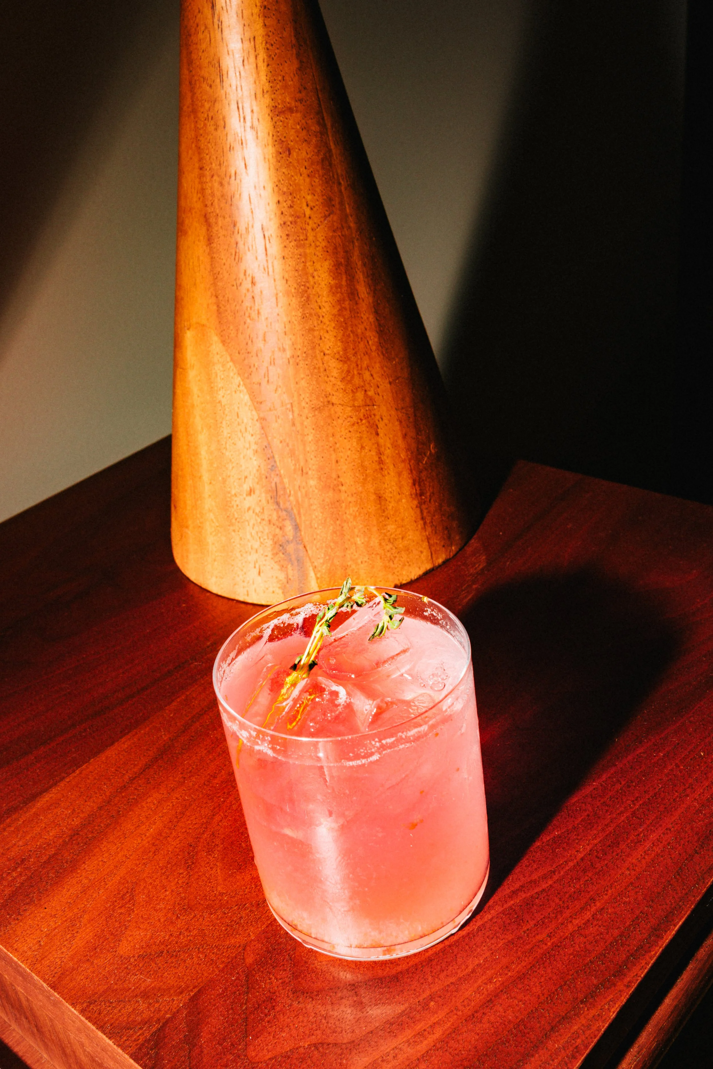 A pink cocktail with ice cubes and a sprig of thyme in a short glass, placed on a wooden surface with a conical wooden object in the background.