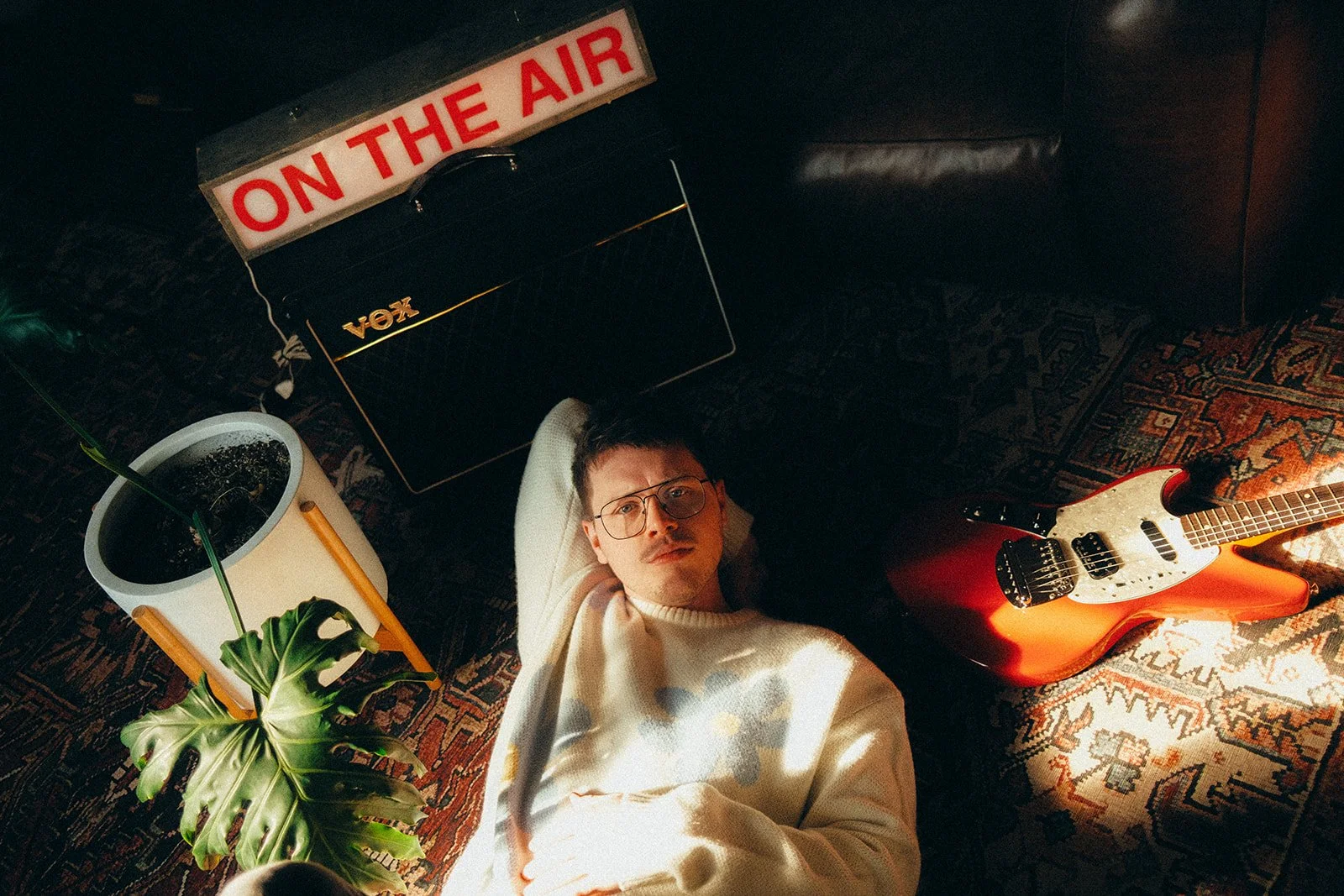 A young man with glasses lying on the floor among plants, a guitar, and a Vox amplifier, with an illuminated 'On The Air' sign above him.