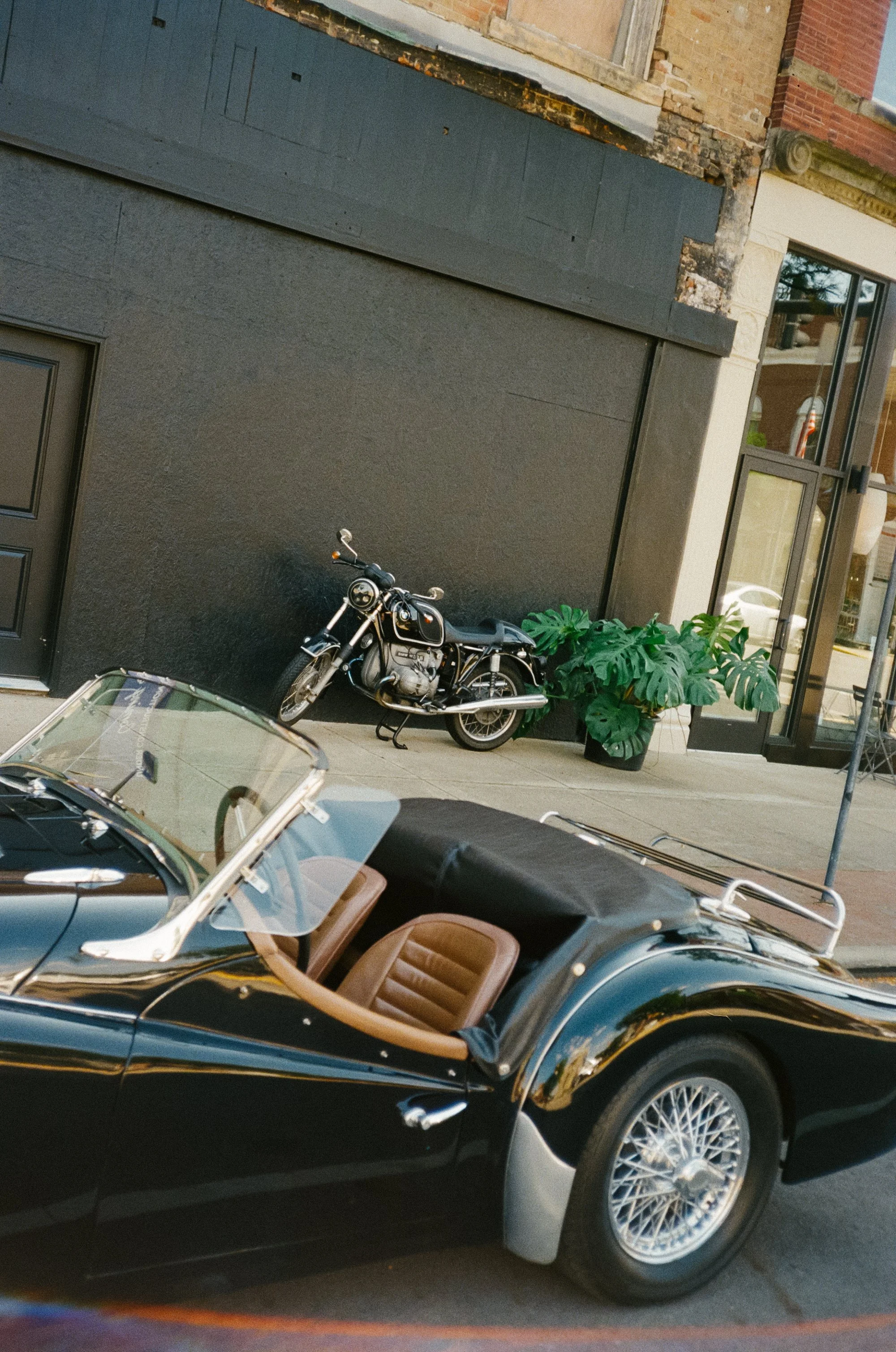 A vintage black convertible sports car parked on the street with tan interior, a classic motorcycle parked against a black building wall, large green potted plants, and a storefront with large glass windows.