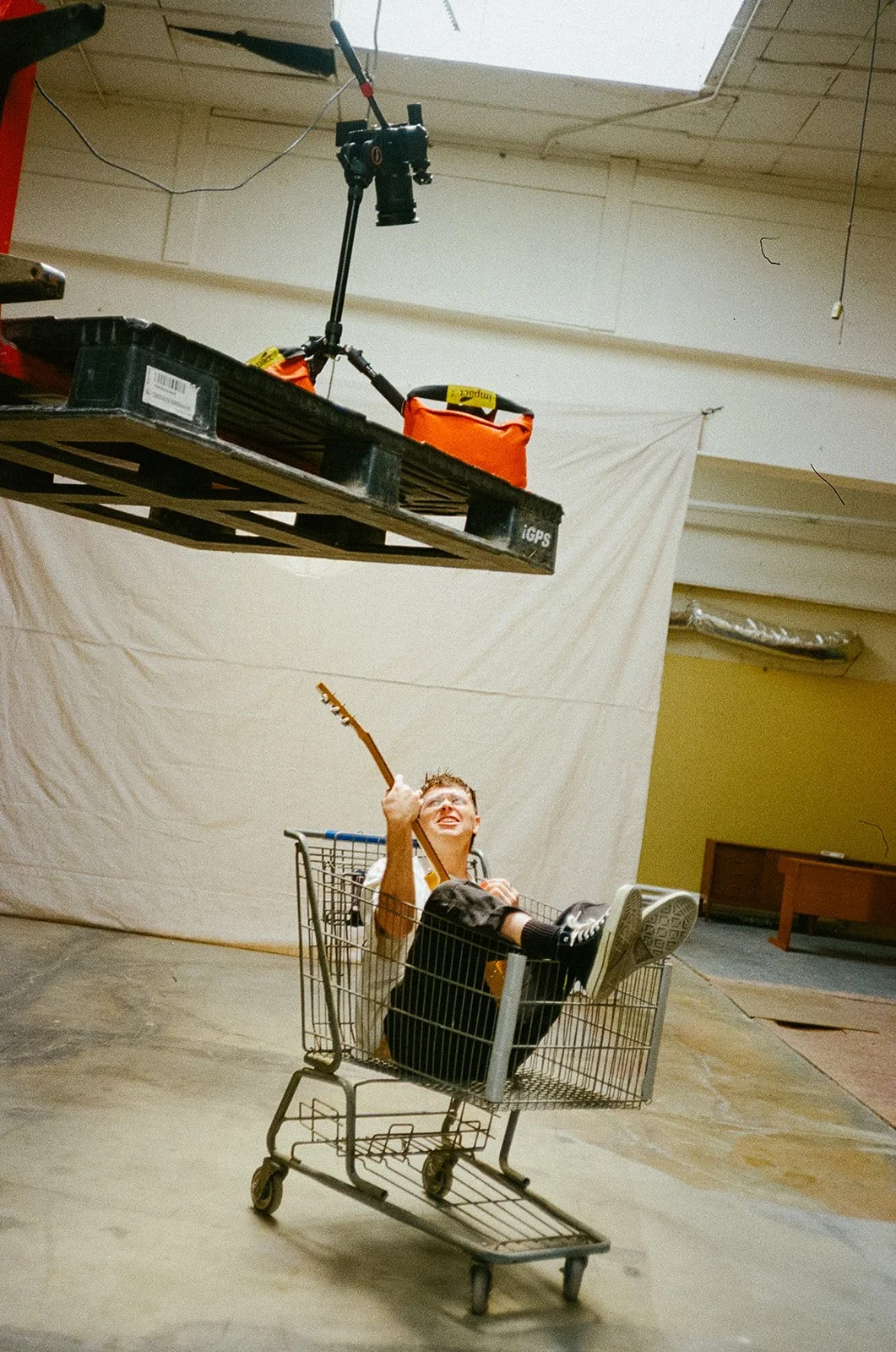 A young person is sitting in a shopping cart, holding a guitar and smiling, with a backdrop of a plain white sheet hanging on a wall