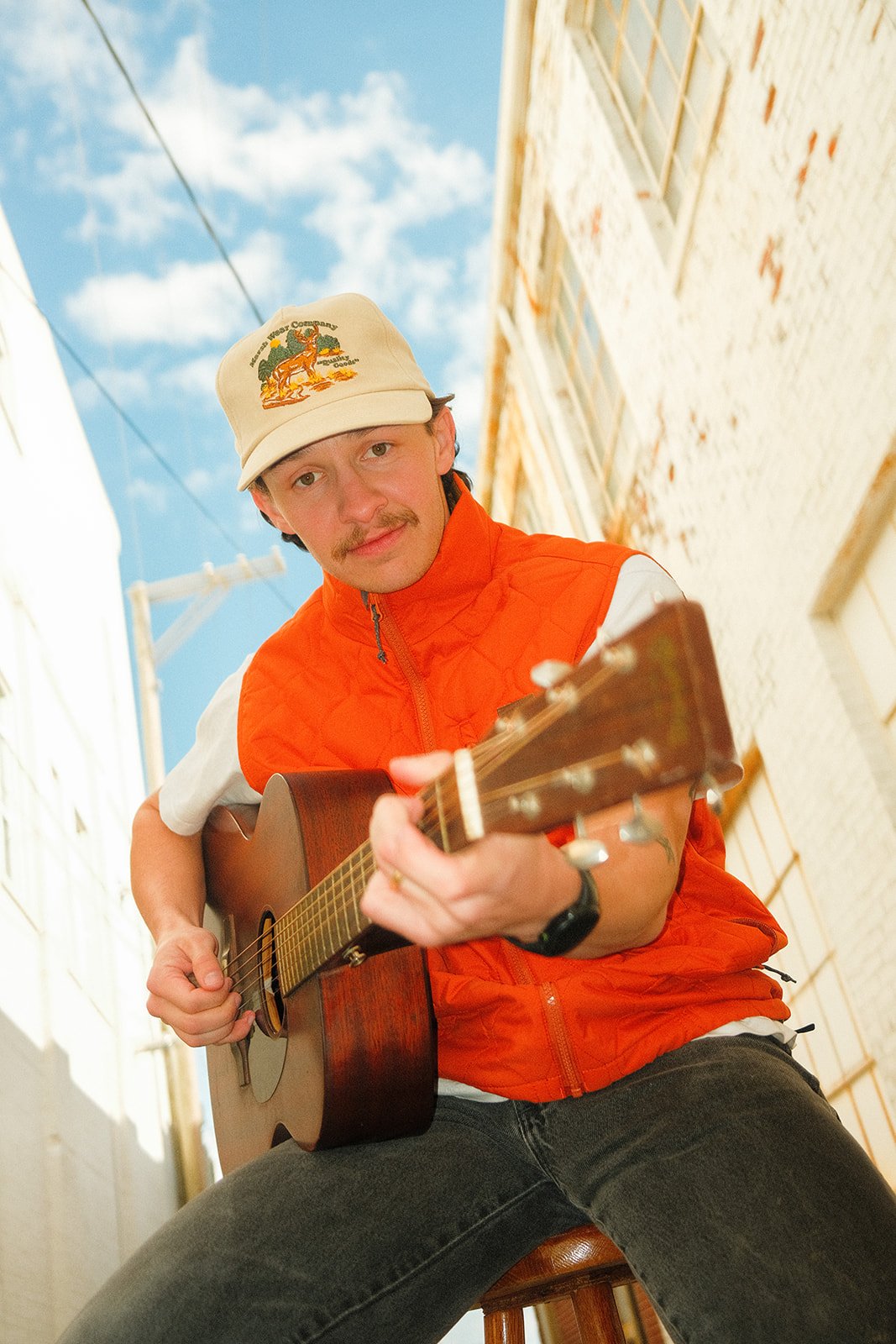 A man wearing an orange vest and a beige cap with a hiking logo, playing an acoustic guitar in an outdoor alley with white brick walls and a blue sky.