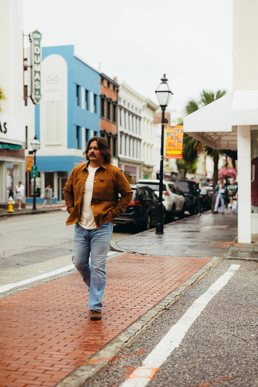 A man with medium-length hair and a mustache walking on a brick crosswalk in a downtown area with colorful buildings, parked cars, and street lamps.