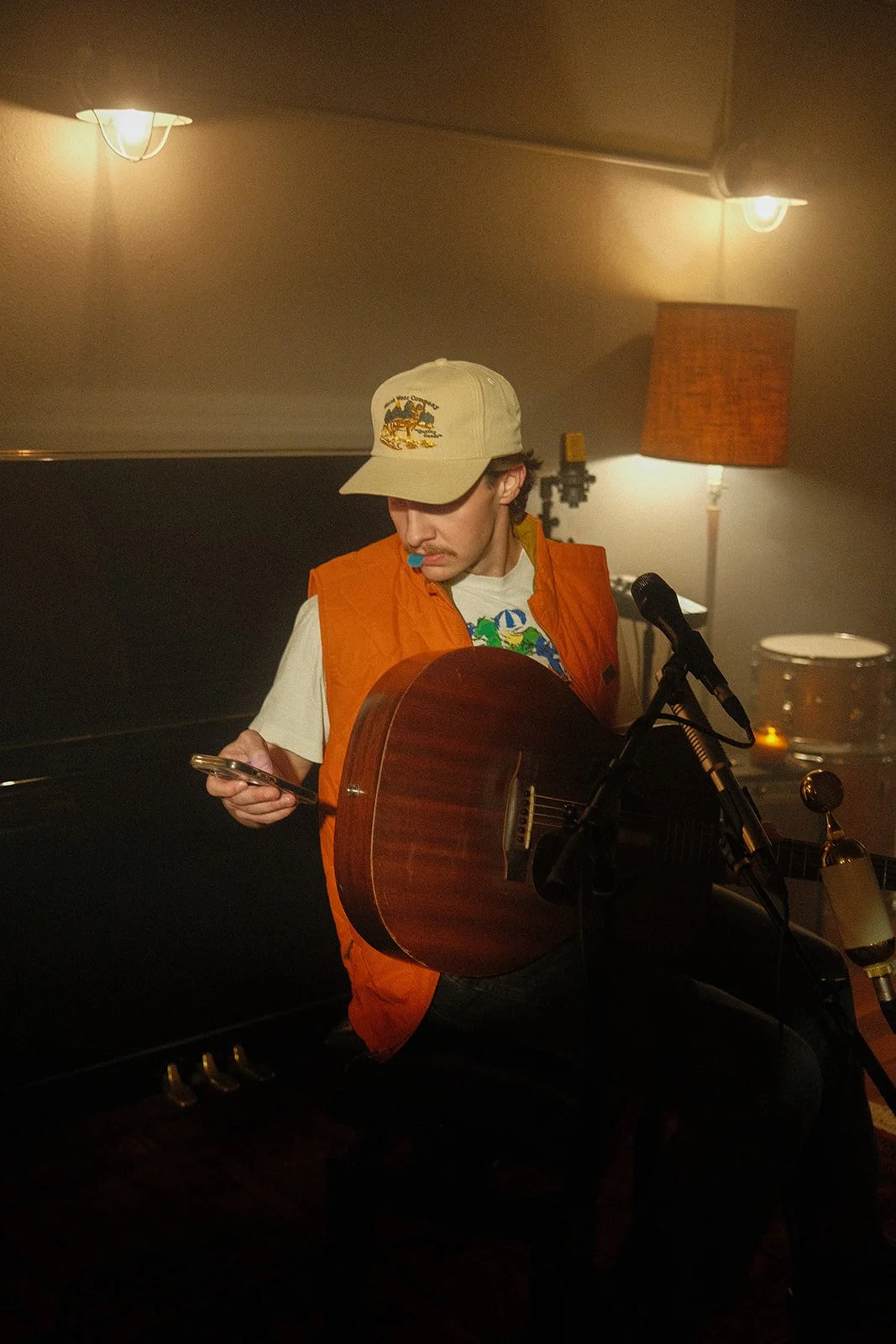 A young man with curly hair and a mustache, wearing a beige baseball cap, orange vest, and a graphic t-shirt, sitting with an acoustic guitar on his lap, looking at his phone in a dimly lit room with a microphone in front of him.
