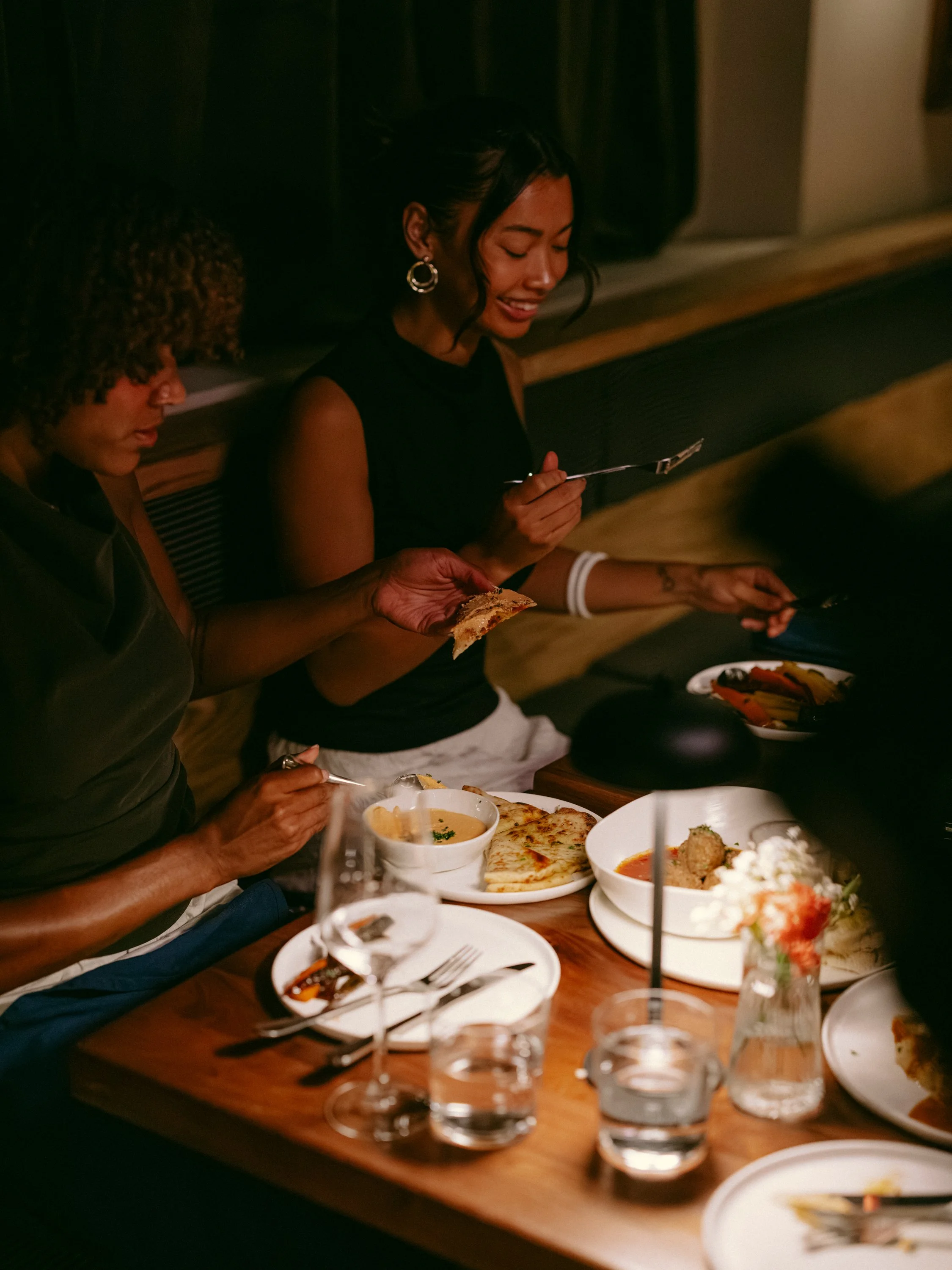 People sitting at a dinner table with plates of food, including pizza, vegetables, and sauces, in a dimly lit restaurant.