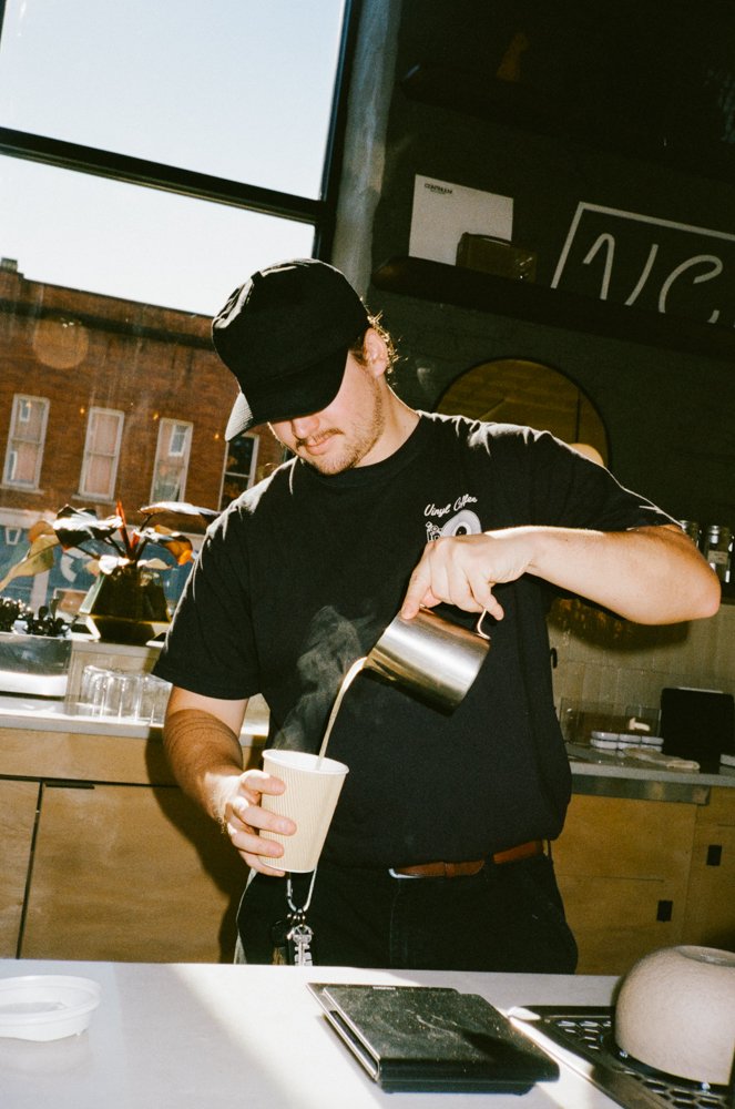 A barista pouring steamed milk into a paper cup at a coffee shop with a large window behind him