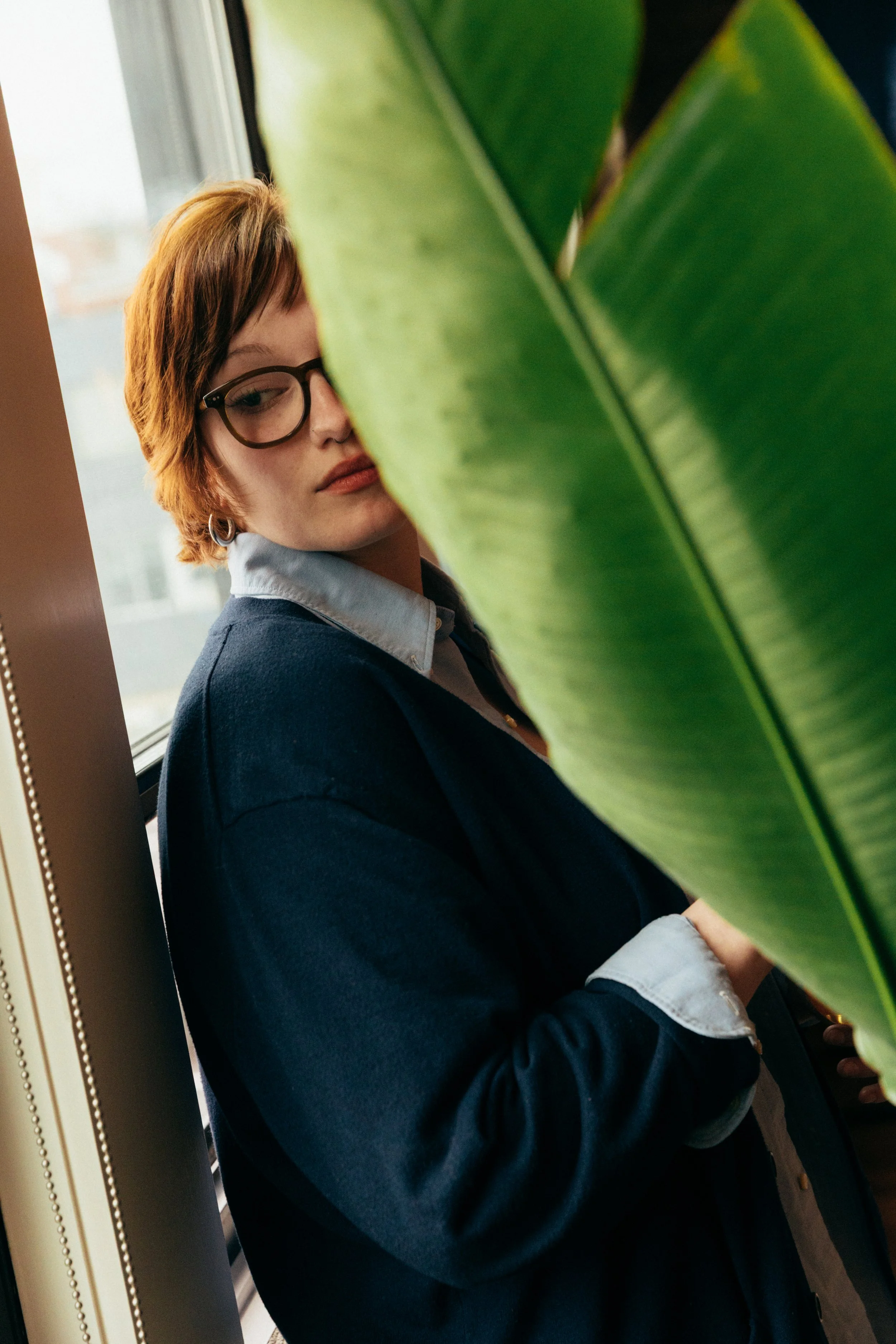 A young woman with short brown hair and glasses, wearing a blue blazer and a light blue shirt, is standing near a window with green leaves partially obscuring her face.