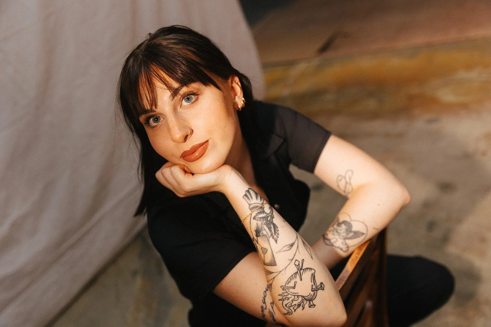 A young woman with dark brown hair and tattoos, resting her chin on her hand, looking at the camera with a slight smile.