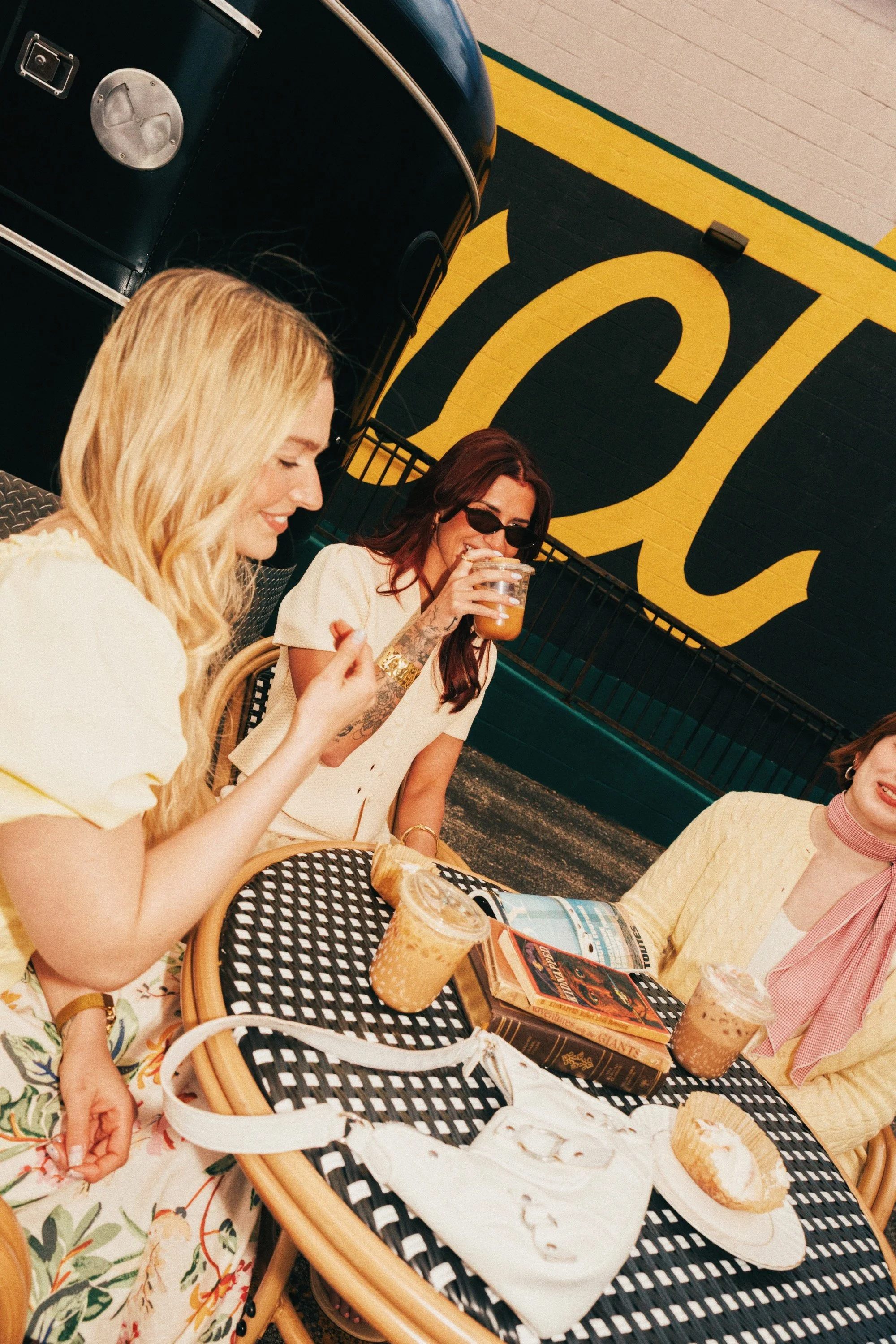 Three women are sitting at a table outside a cafe, enjoying drinks. The table has ice cream and a latte. The background features a black wall with a large yellow 
