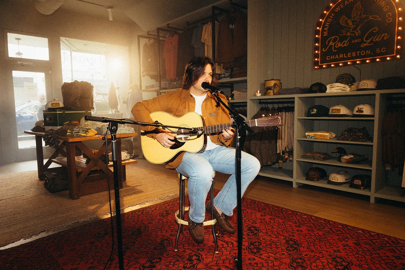 A person with shoulder-length hair, wearing a yellow jacket, white shirt, and jeans, is sitting on a stool playing an acoustic guitar and singing into a microphone inside a retail store that sells hats, apparel, and accessories. The store has warm li