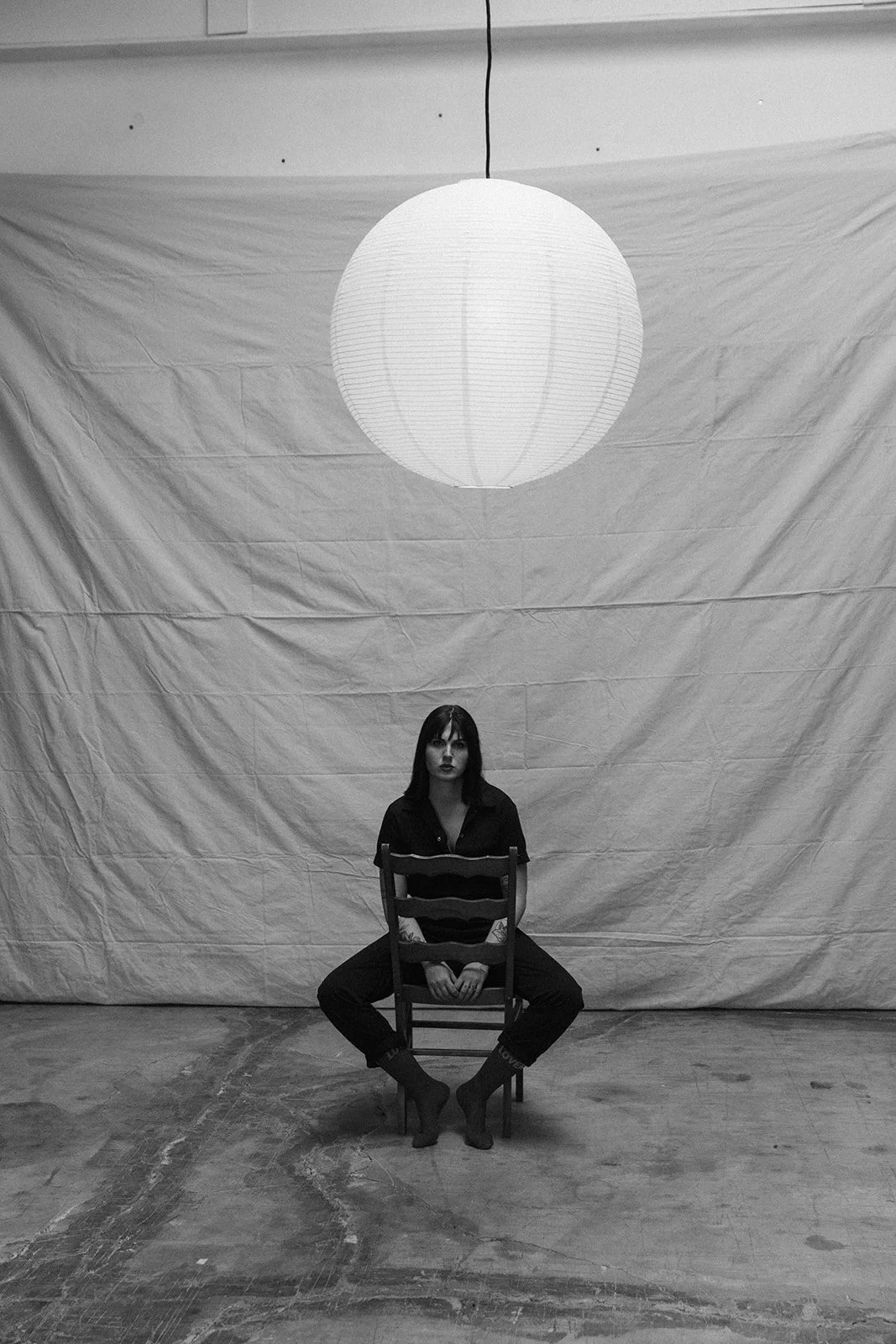 A woman with dark hair and dark clothing sits on a wooden chair in a room with a plain backdrop and a large paper lantern hanging from the ceiling.