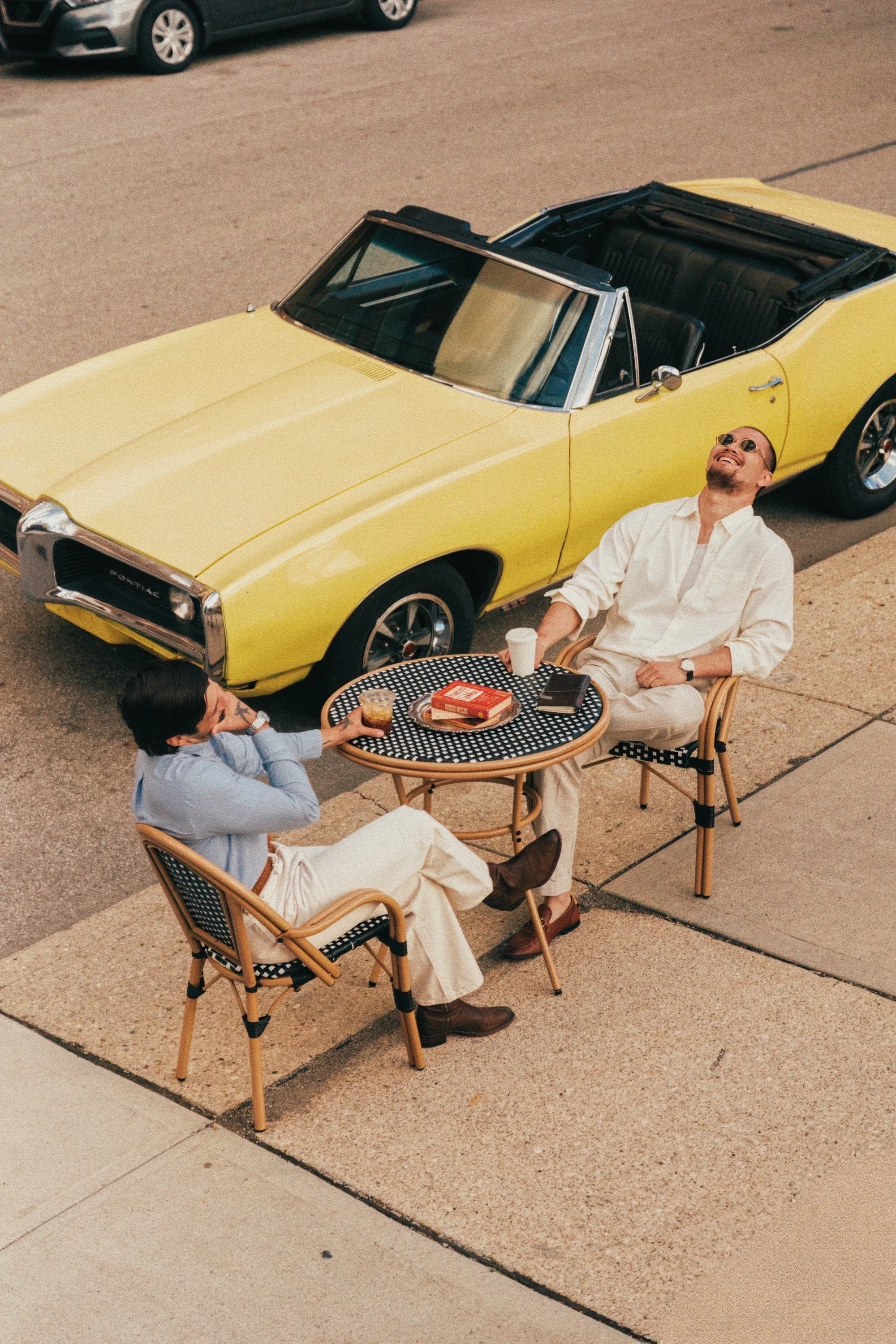 Two men sitting outdoors at a round table with drinks, with a yellow convertible car parked nearby; one man is wearing sunglasses, a white shirt, and beige pants, the other is wearing glasses and a blue shirt. Cafe Stai Manso
