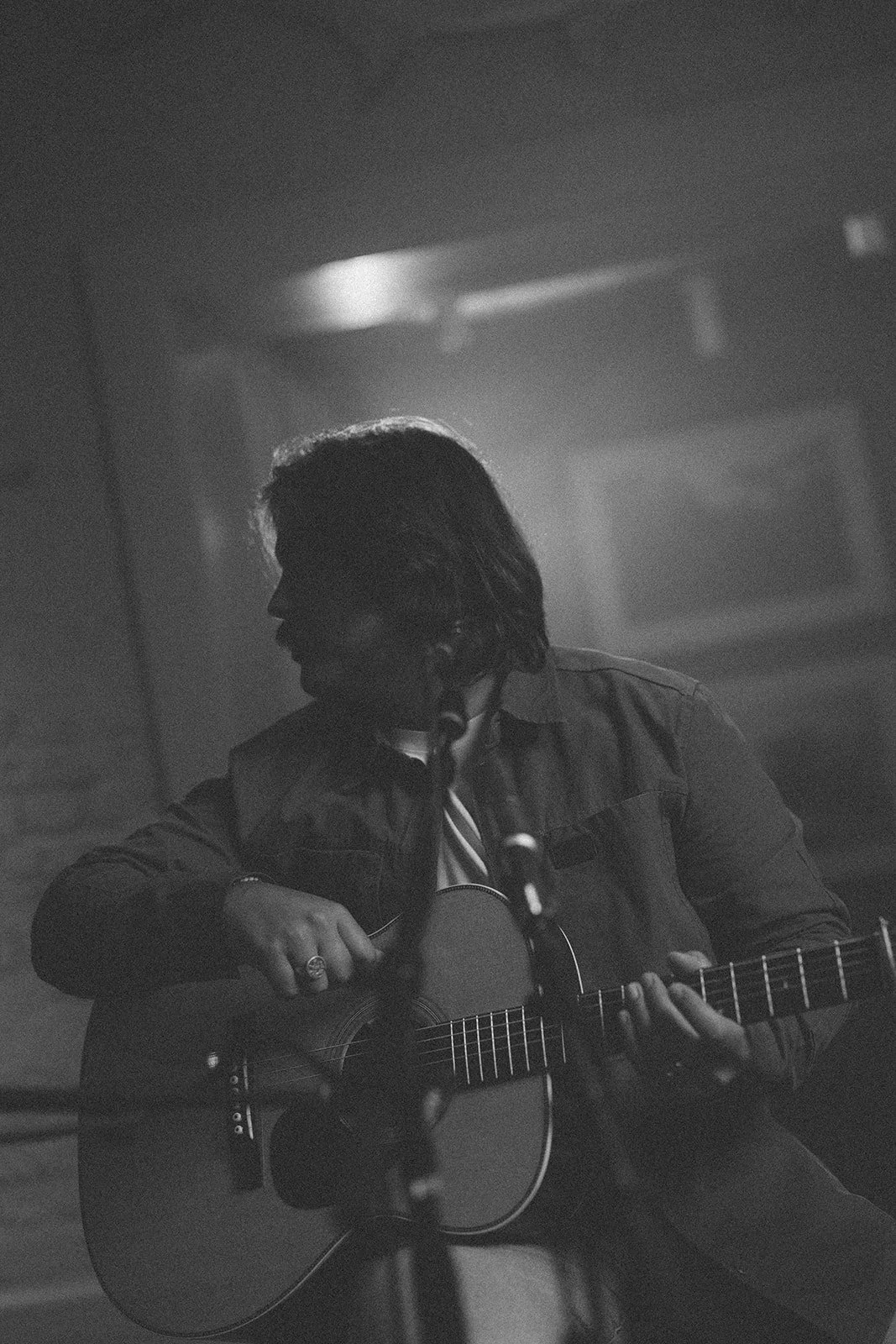 A person with shoulder-length hair playing an acoustic guitar in a dimly lit setting, appears to be performing or practicing, with a microphone in front.
