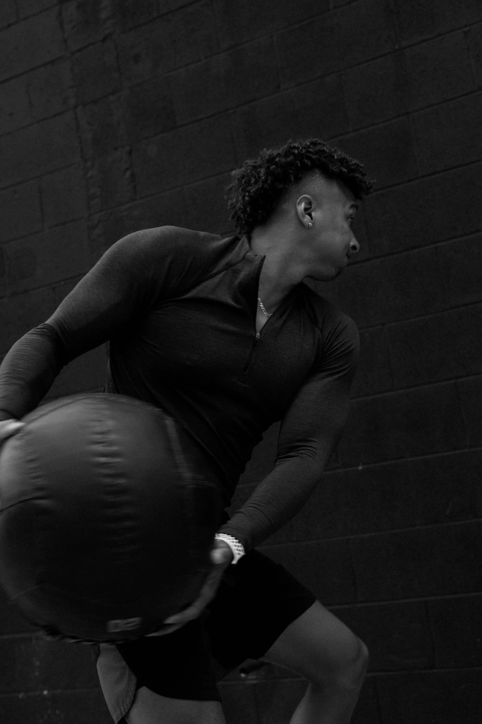 A person with curly hair wearing a long-sleeve shirt and shorts sitting on the ground against a brick wall, partially turned away from the camera.