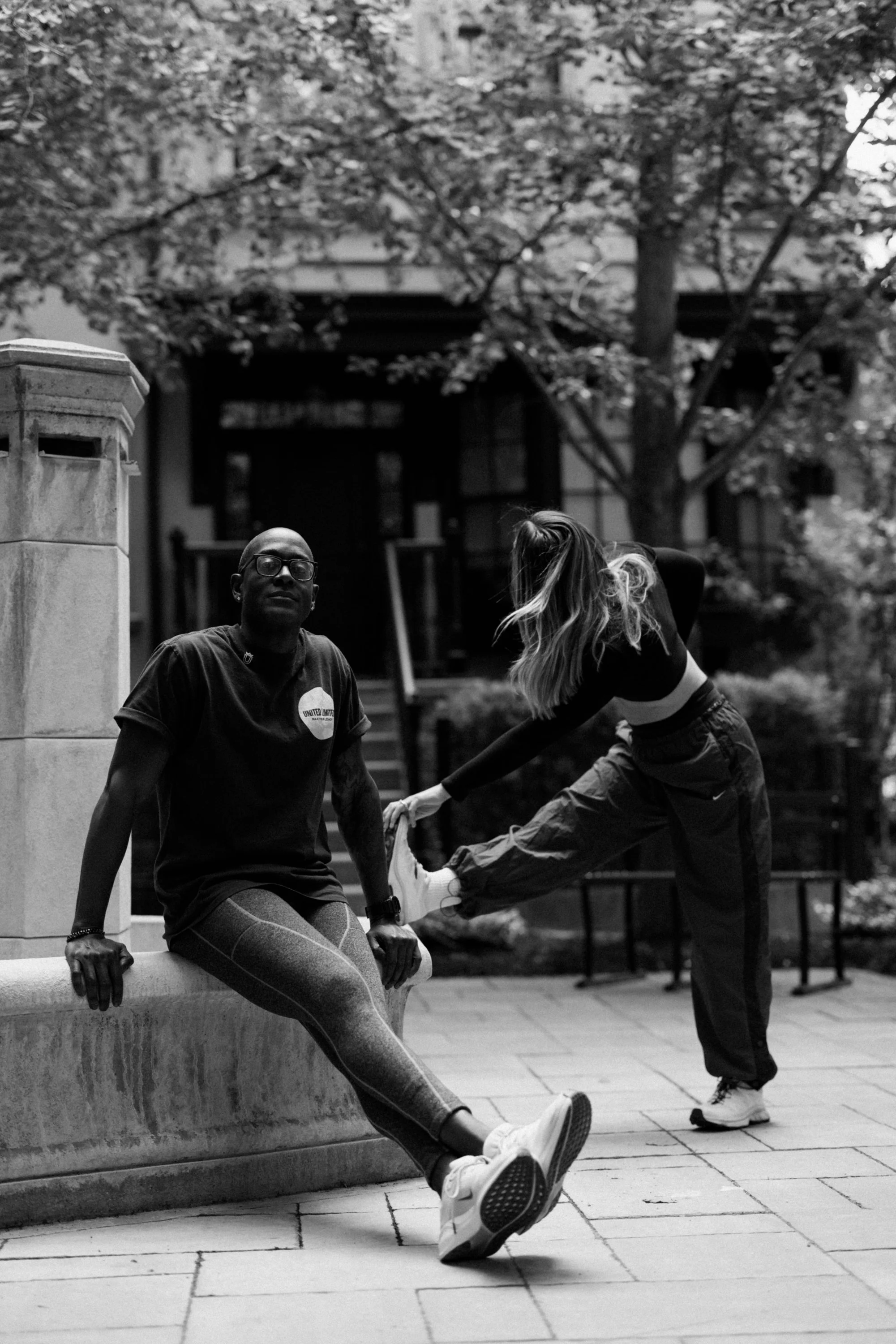 Two women stretching outdoors in front of a house with a tree in the background. One woman is sitting on a stone bench, while the other woman, wearing a tracksuit, is stretching her leg.