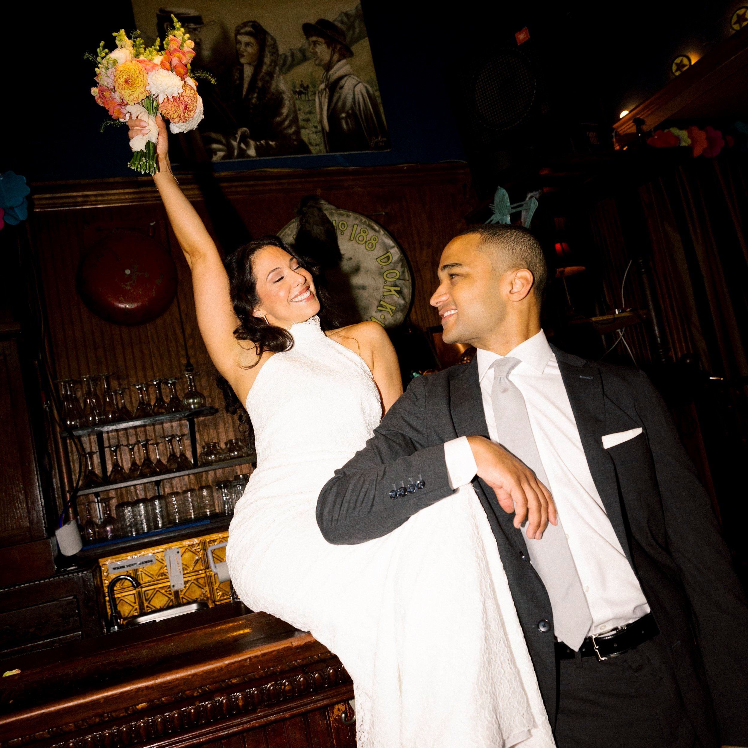A woman in a white dress holding a bouquet of flowers on her shoulder, smiling and dancing with a man in a black suit and white shirt, in a festive indoor setting.